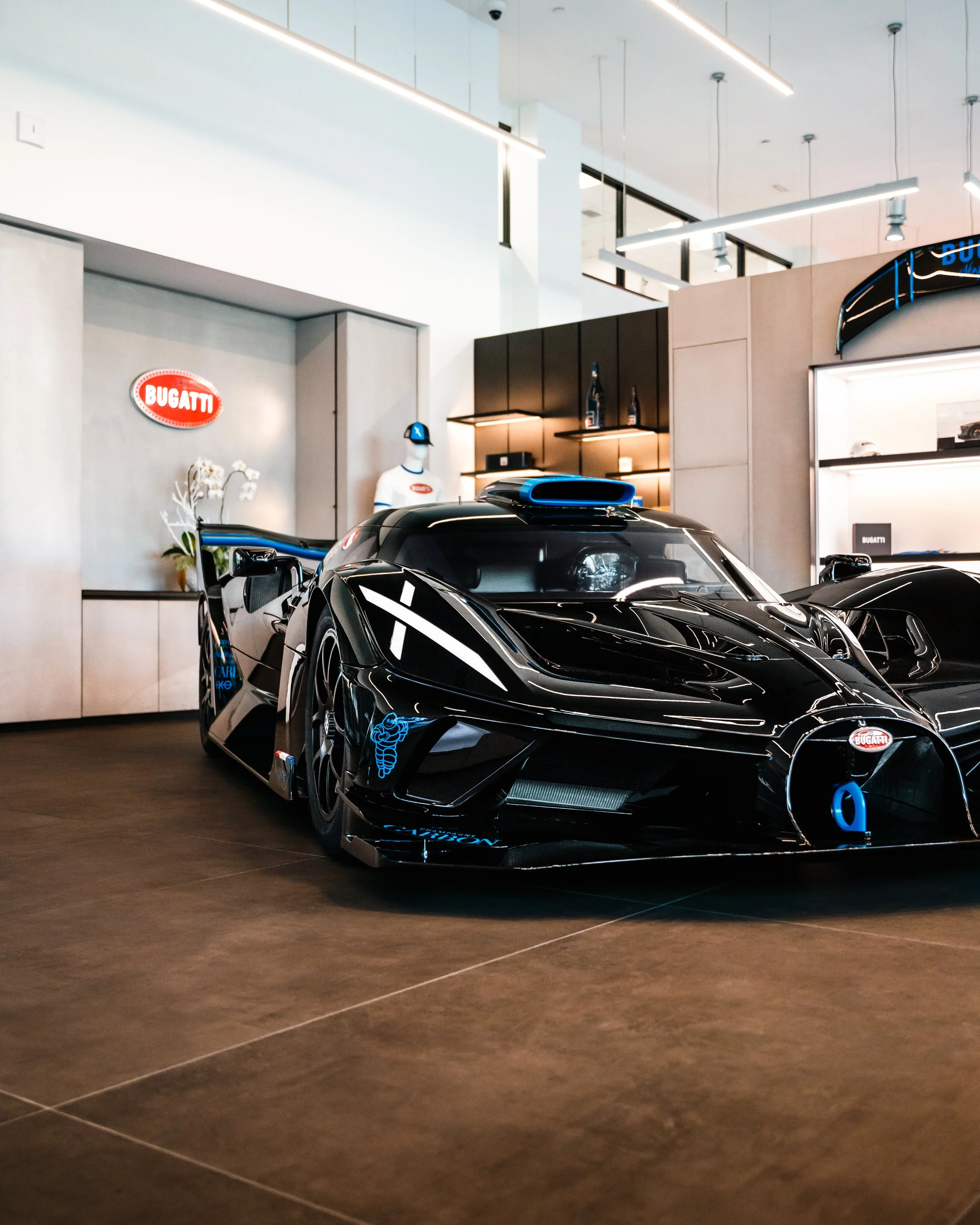A black Bugatti Bolide with blue accents displayed indoors at a showroom, with a Bugatti logo visible on a wall behind it.