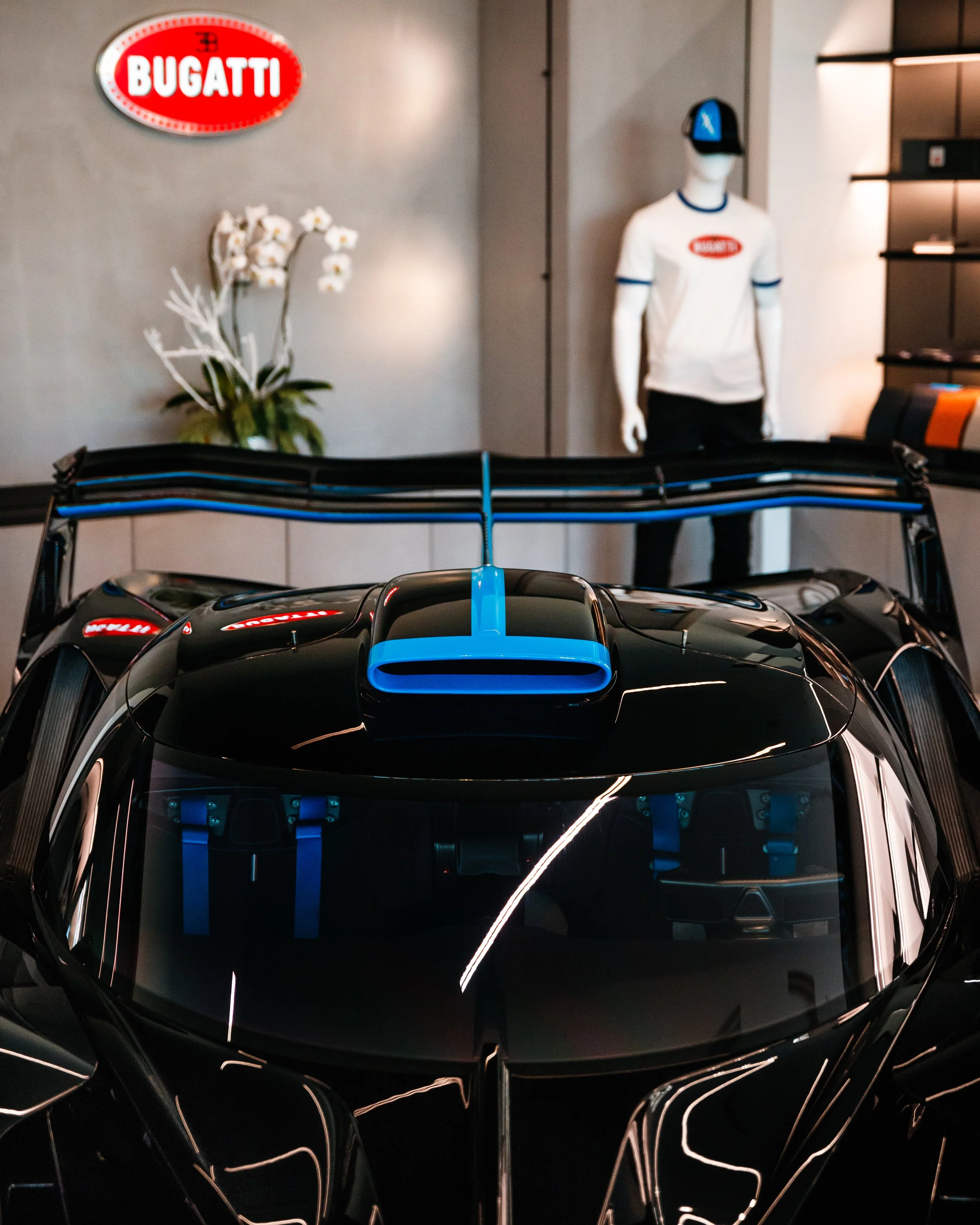 A black Bugatti Bolide with blue accents, viewed from the front, inside a showroom with a Bugatti logo sign, mannequin wearing a Bugatti T-shirt and cap, and a white orchid plant in the background.