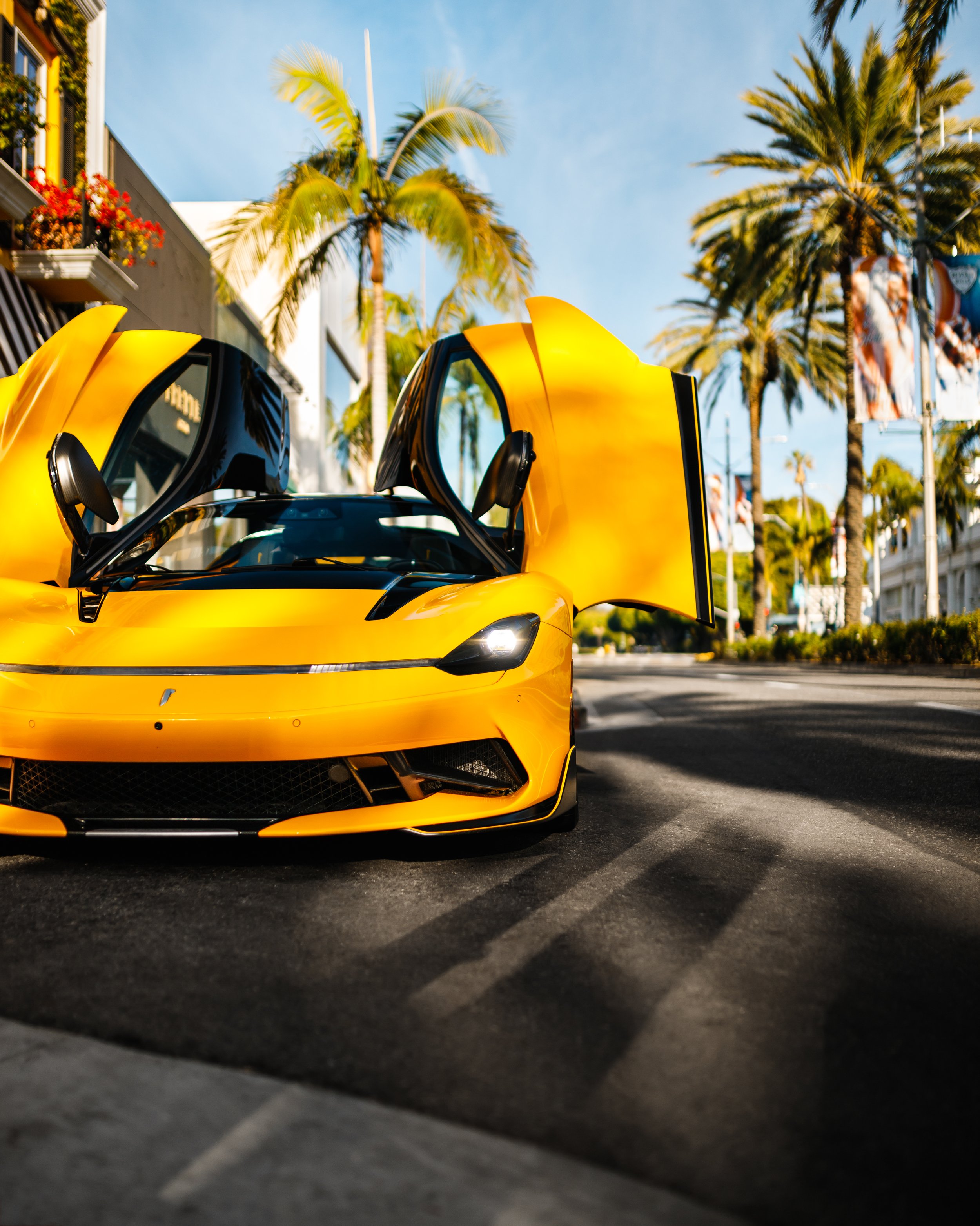 Yellow Pininfarina Battista car with open gull-wing doors parked on a palm tree-lined street