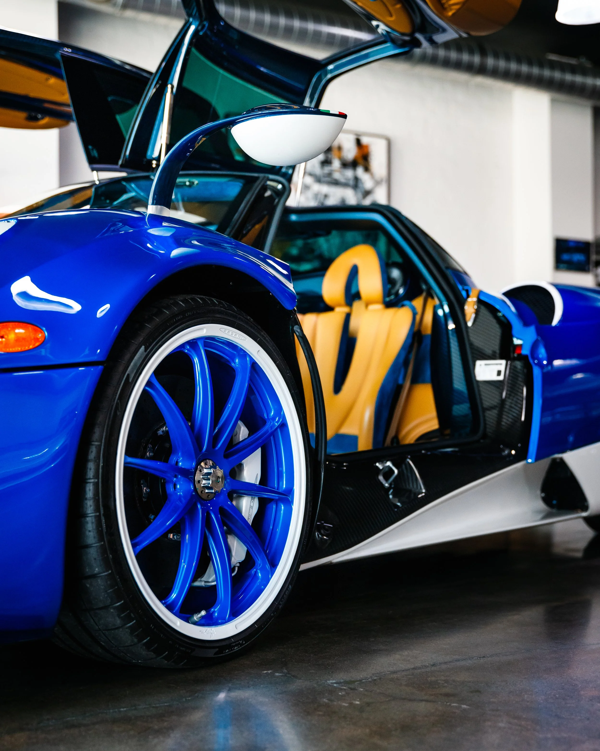 A close-up of a blue Pagani Huayra with an open gullwing door, showing the tan interior and the front wheel with a matching blue rim.