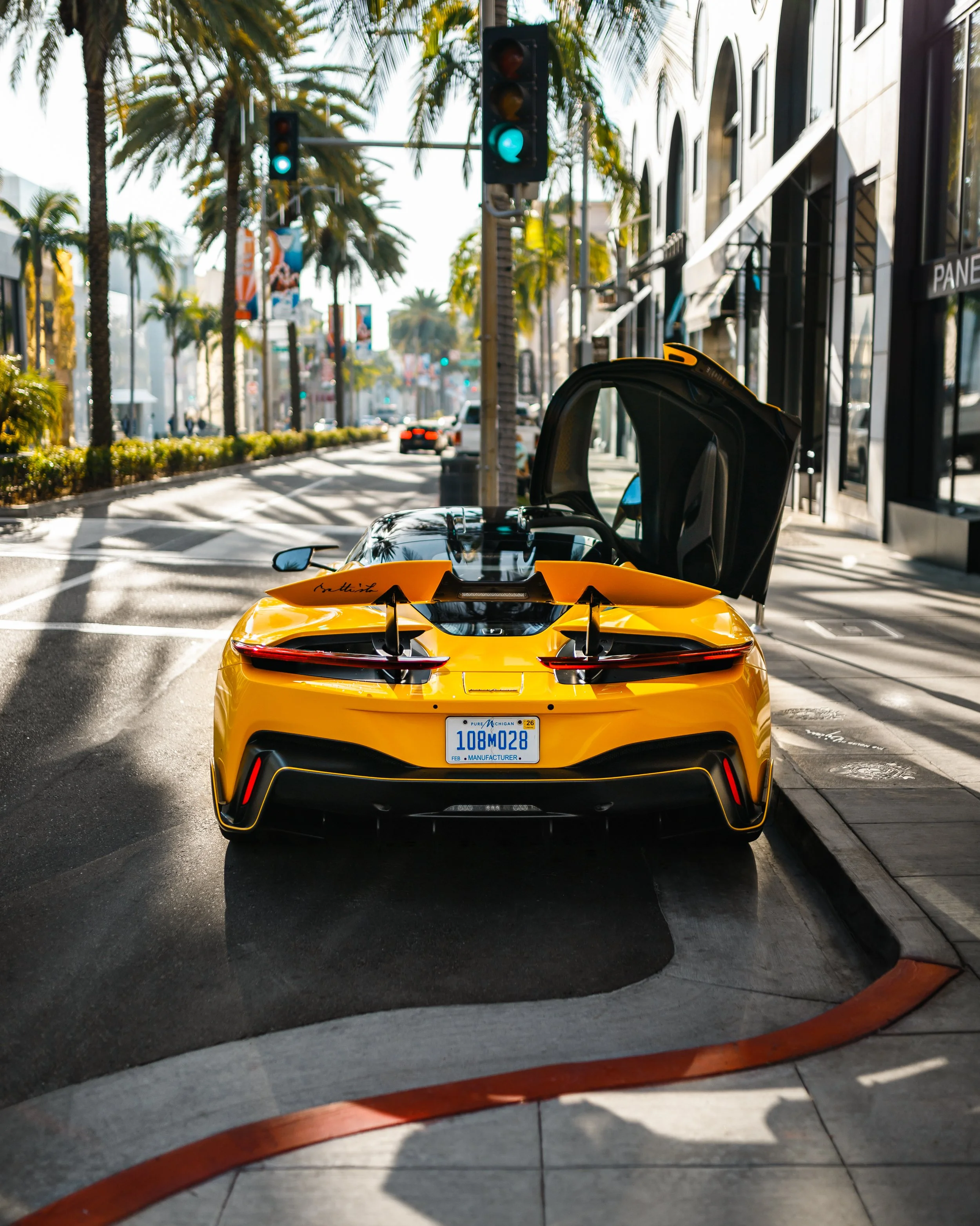 A yellow Pininfarina Battista with butterfly doors open parked on a city street with palm trees and buildings.