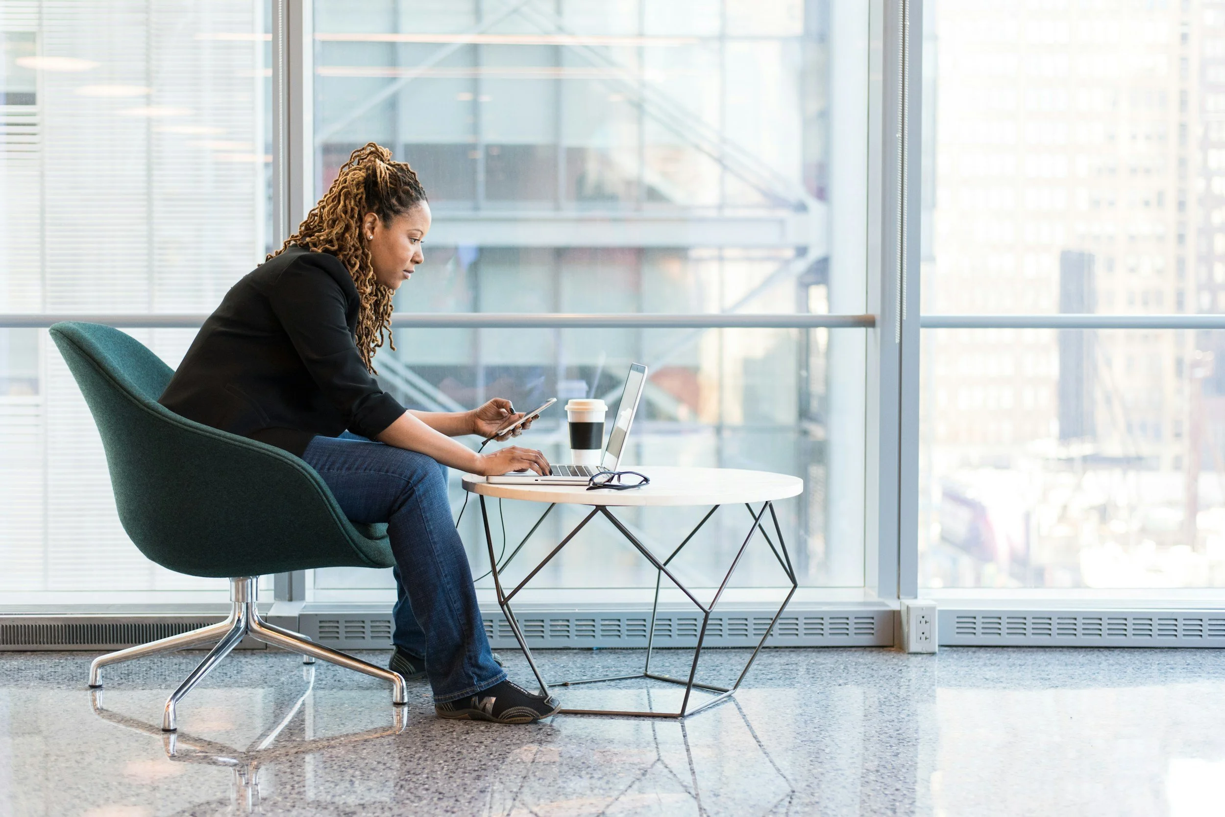 A woman with dreadlocks sitting in a modern office lounge, looking at her smartphone with a laptop and coffee on a small table nearby, in front of large windows with a cityscape view.