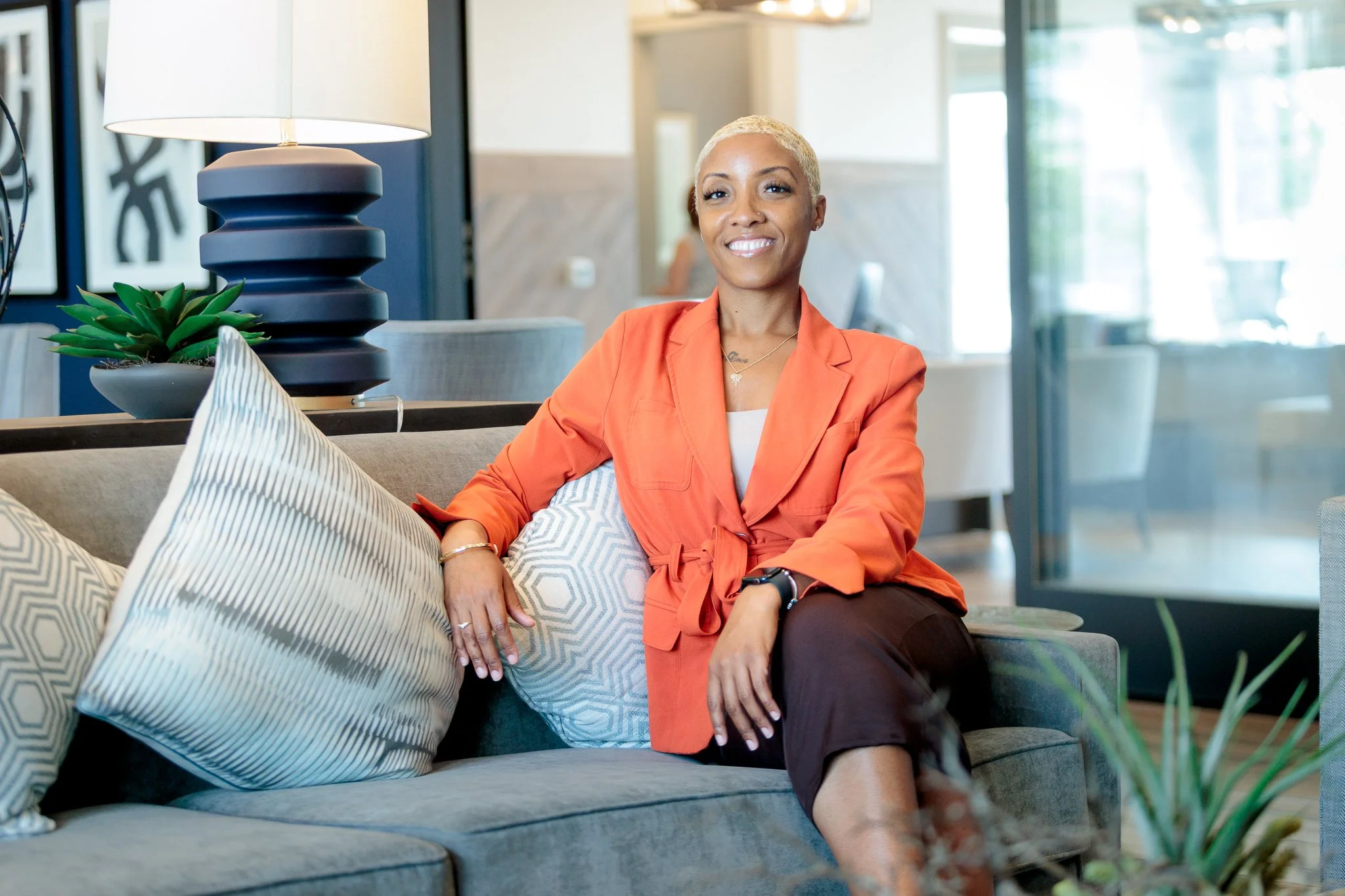 A woman with short blonde hair and a bright smile, sitting on a gray sofa in a modern, well-lit room, wearing an orange blazer and dark pants.