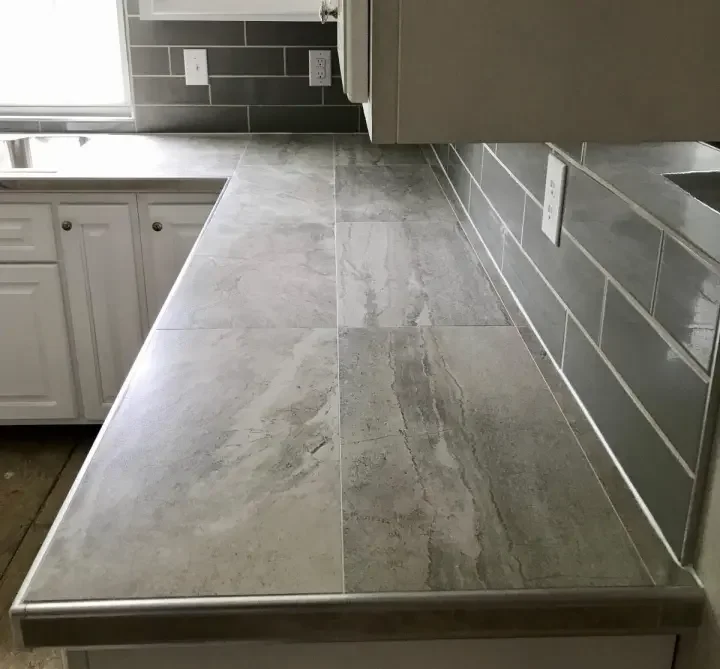Close-up of a kitchen countertop with large gray stone tiles, gray backsplash, and white cabinets in the background.