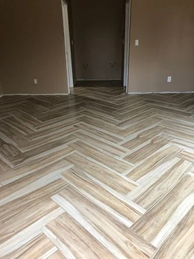 Empty room with wood-patterned herringbone flooring and beige walls.