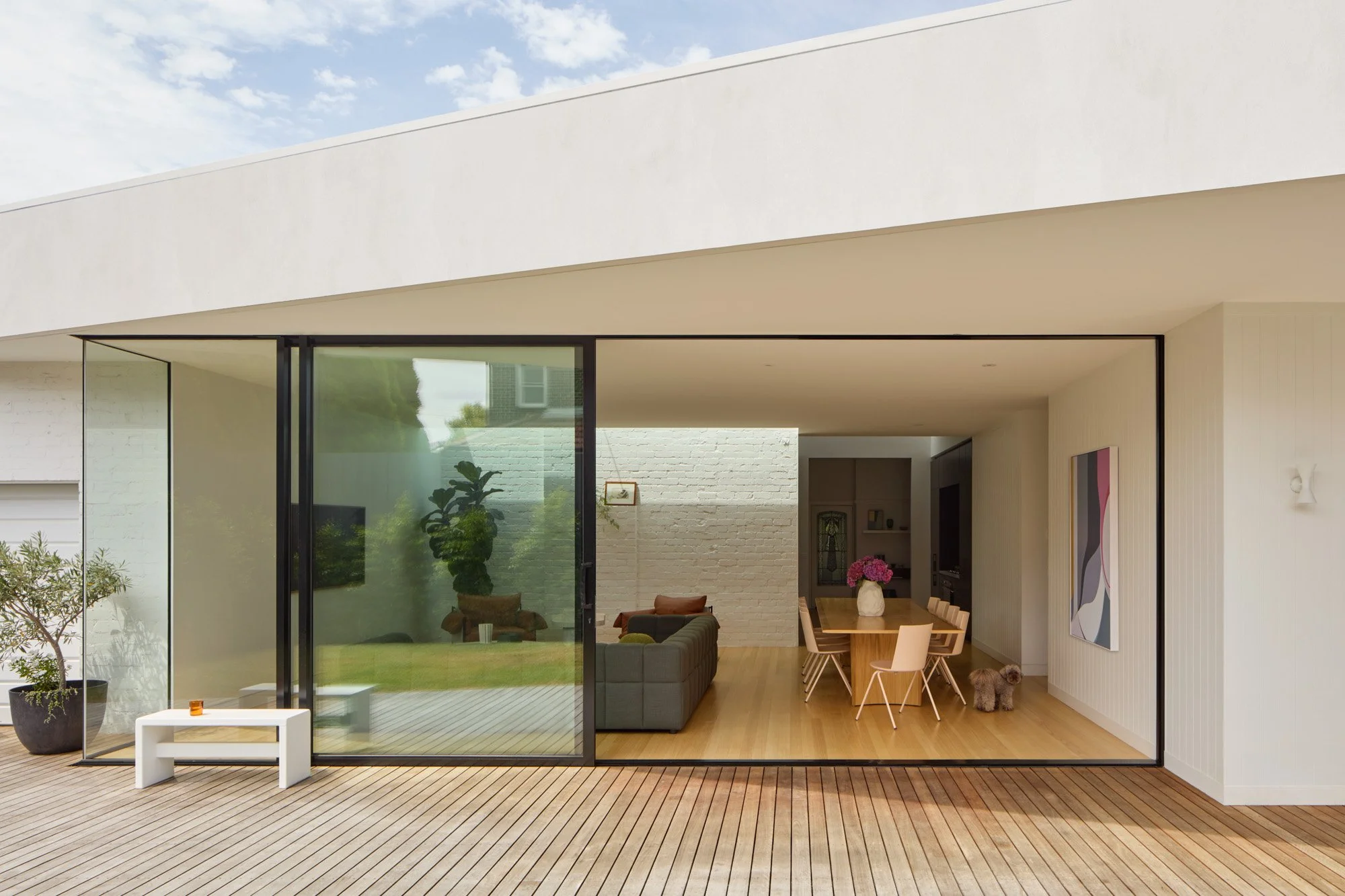 Modern house with large glass sliding doors opening to a wooden deck, with plants and indoor furniture visible inside.