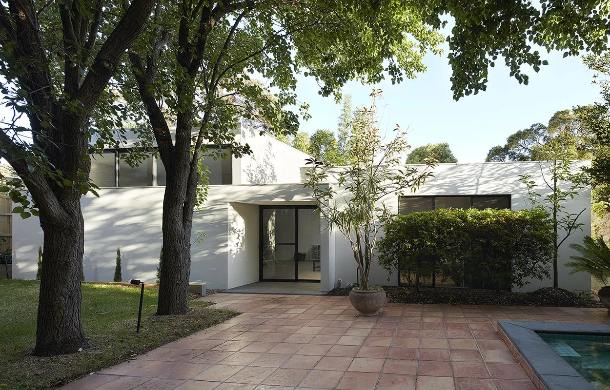 Modern white house with large glass door, surrounded by trees and greenery, with a tiled patio and a small pool on the right.