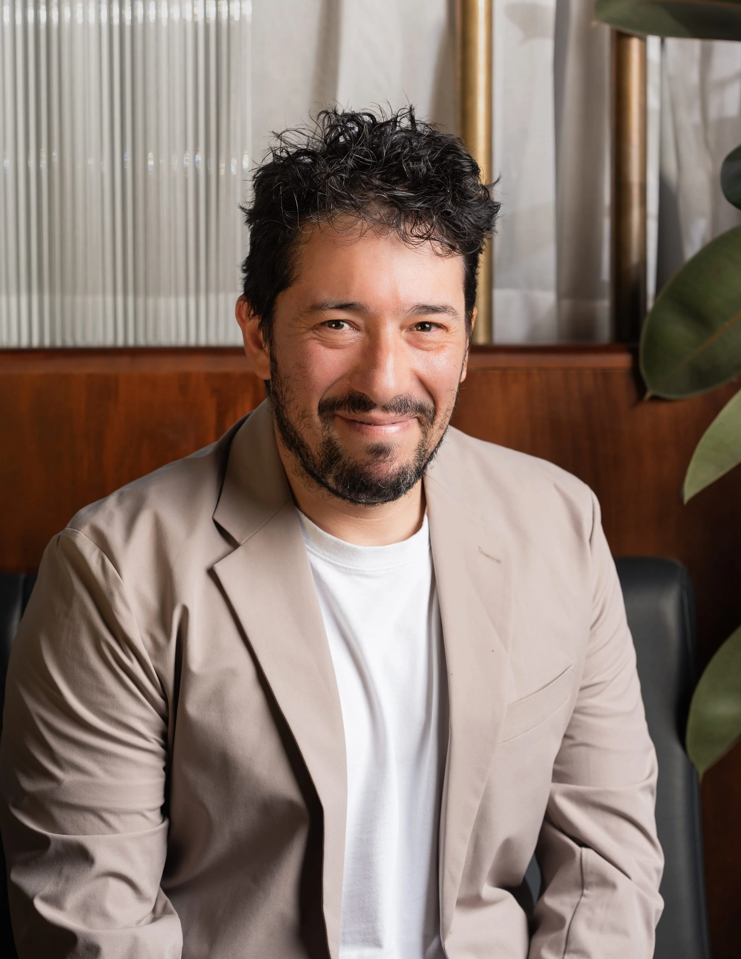 A man with dark curly hair and a beard, wearing a beige blazer over a white shirt, smiles at the camera indoors. Behind him are a wood panel, a vertical metallic divider, and some greenery.