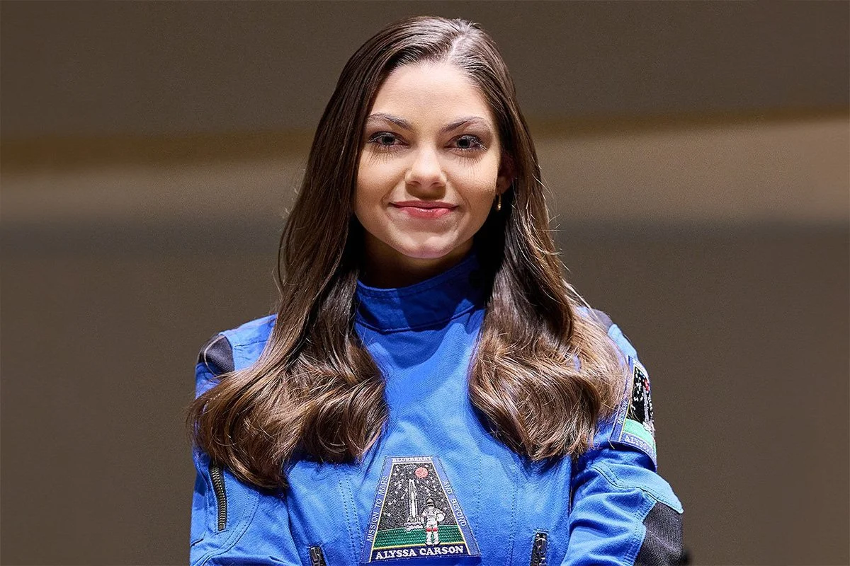Alyssa Carson, a young woman with brown hair, wearing a blue space suit with patches, standing and smiling.