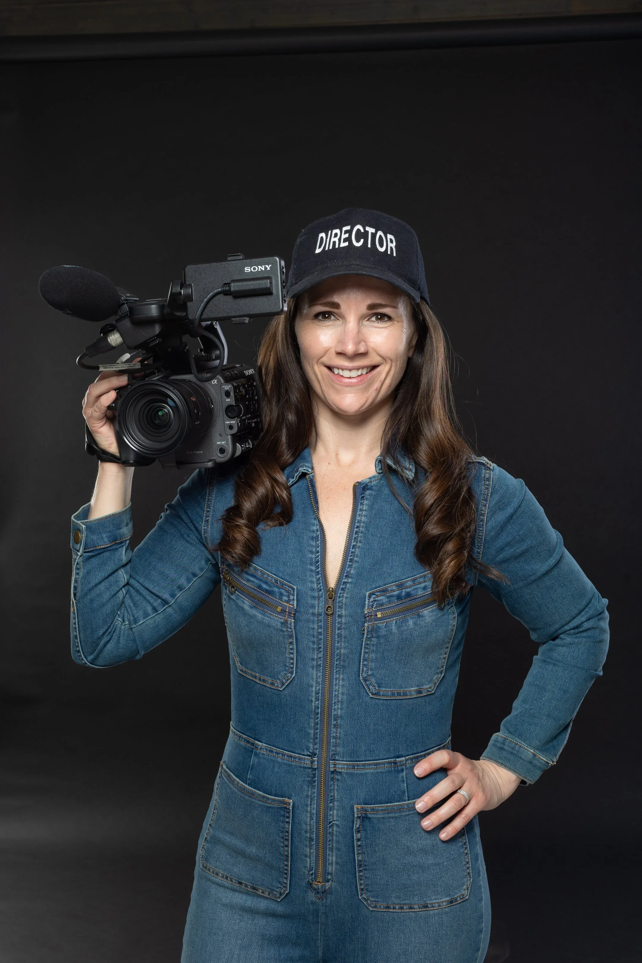 Sarah Moshman holds a professional video camera over her shoulder, wearing a denim jumpsuit and a black cap labeled 'DIRECTOR,' smiling against a black background.