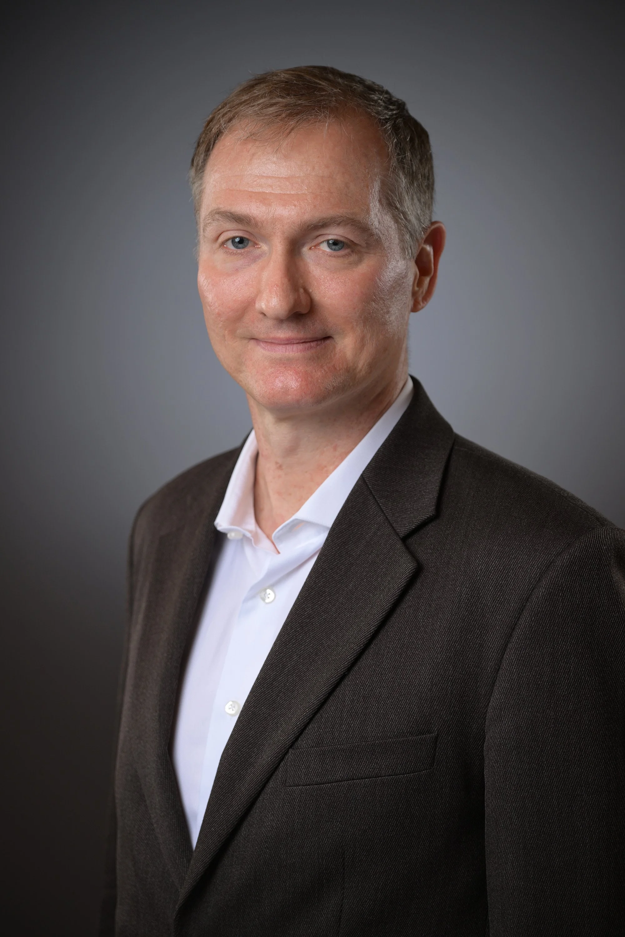 Professional portrait of a man in a dark suit and white shirt, standing against a gray background.