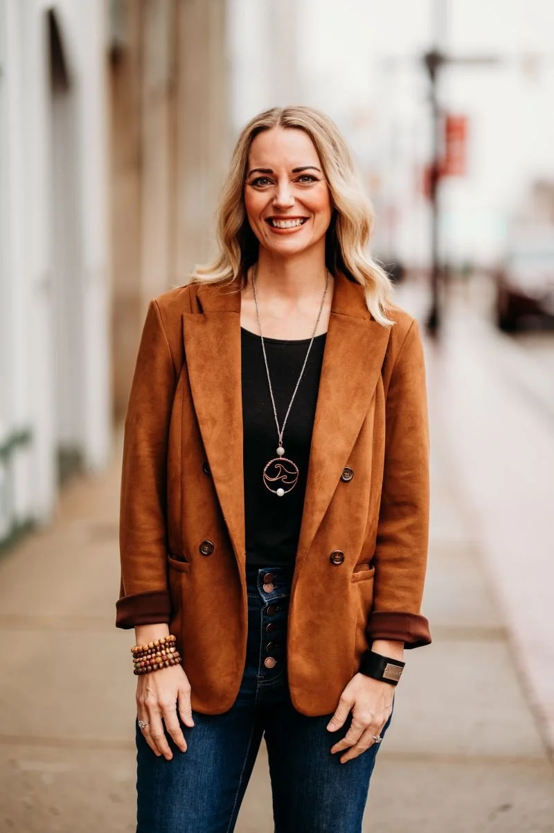 A woman with blonde hair smiling, wearing a brown blazer, black top, jeans, and jewelry, standing on a sidewalk in an urban area.