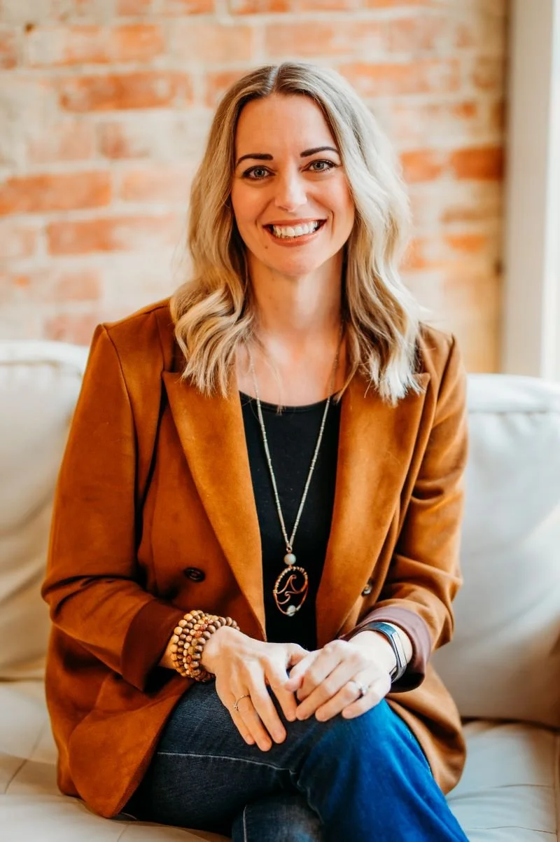 A smiling woman with shoulder-length blonde hair, wearing a brown blazer, black top, and blue jeans, sitting on a beige couch in front of a brick wall.