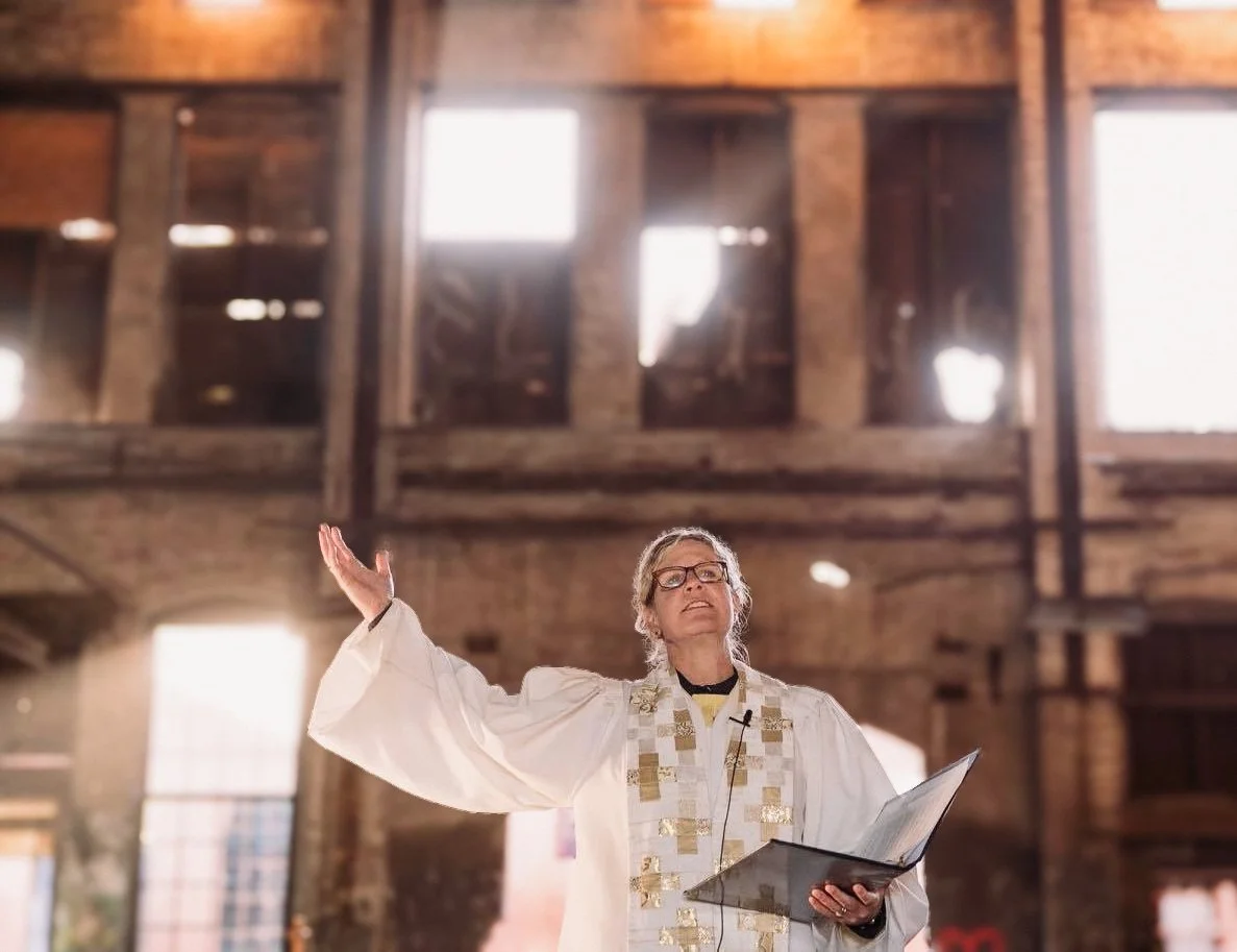 A woman in a white religious robe reading from a book and gesturing with her right hand inside a rustic church.