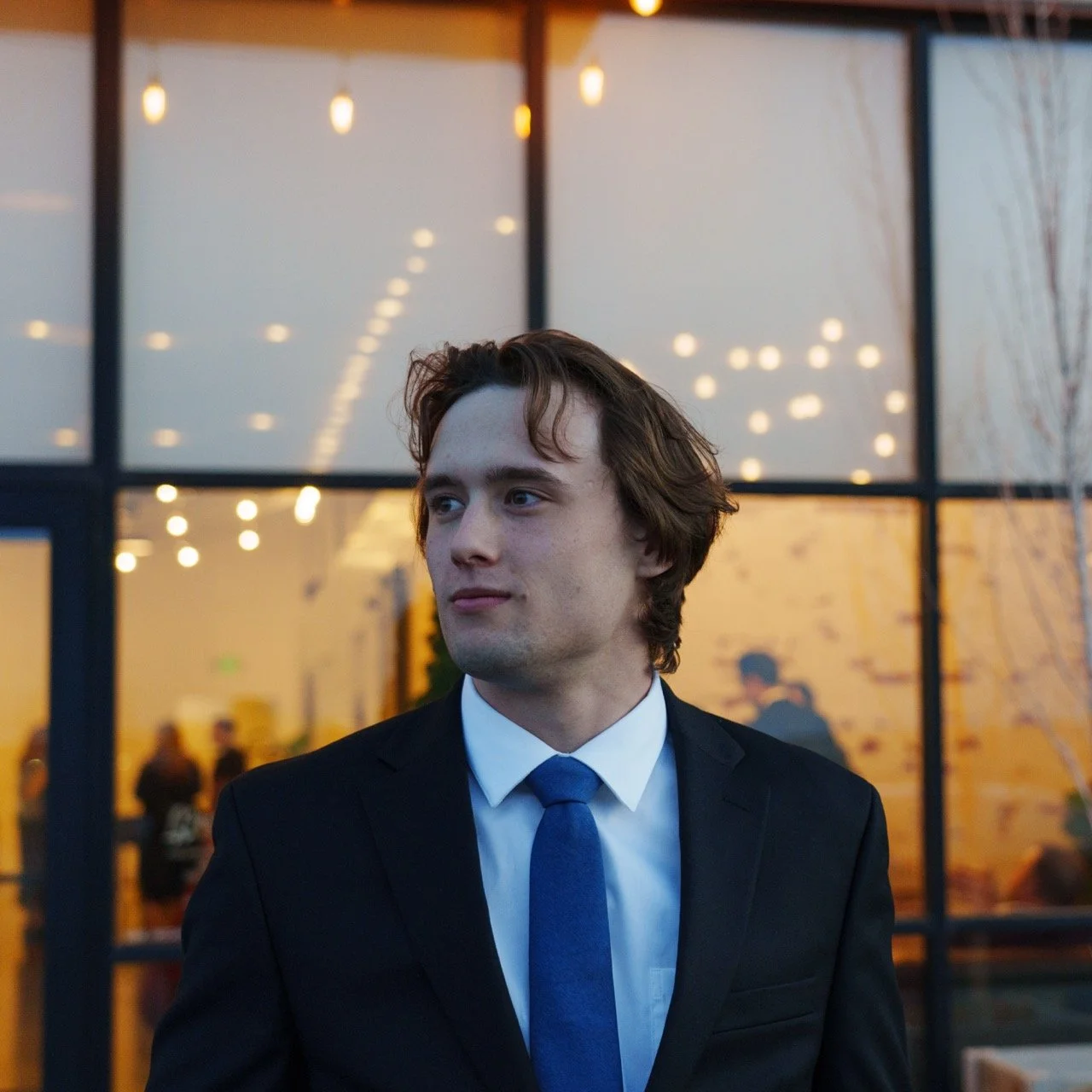 A young man in a black suit and blue tie standing outdoors, looking to the side, with a building and warm lights in the background.