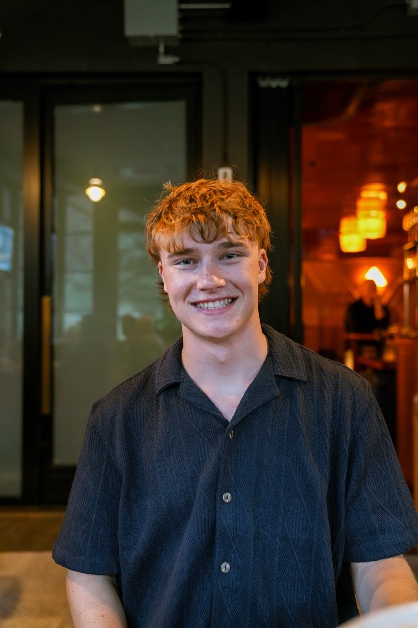 A young man with curly red hair and a big smile, wearing a black collared shirt, standing inside a warmly lit restaurant or cafe.