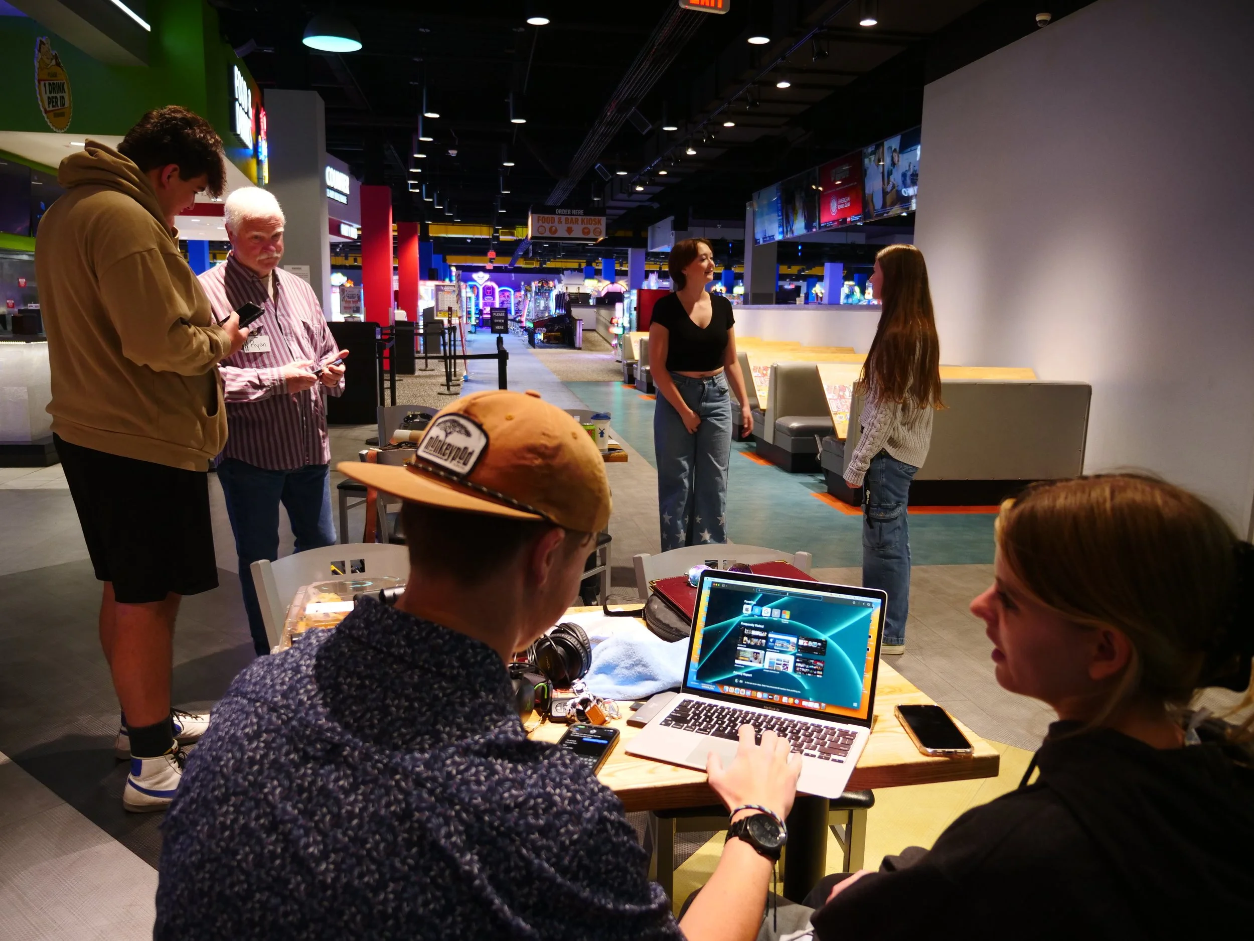 People gathered around a table with laptops, smartphones, and a computer screen in an arcade or gaming area with bright lights and arcade machines in the background.