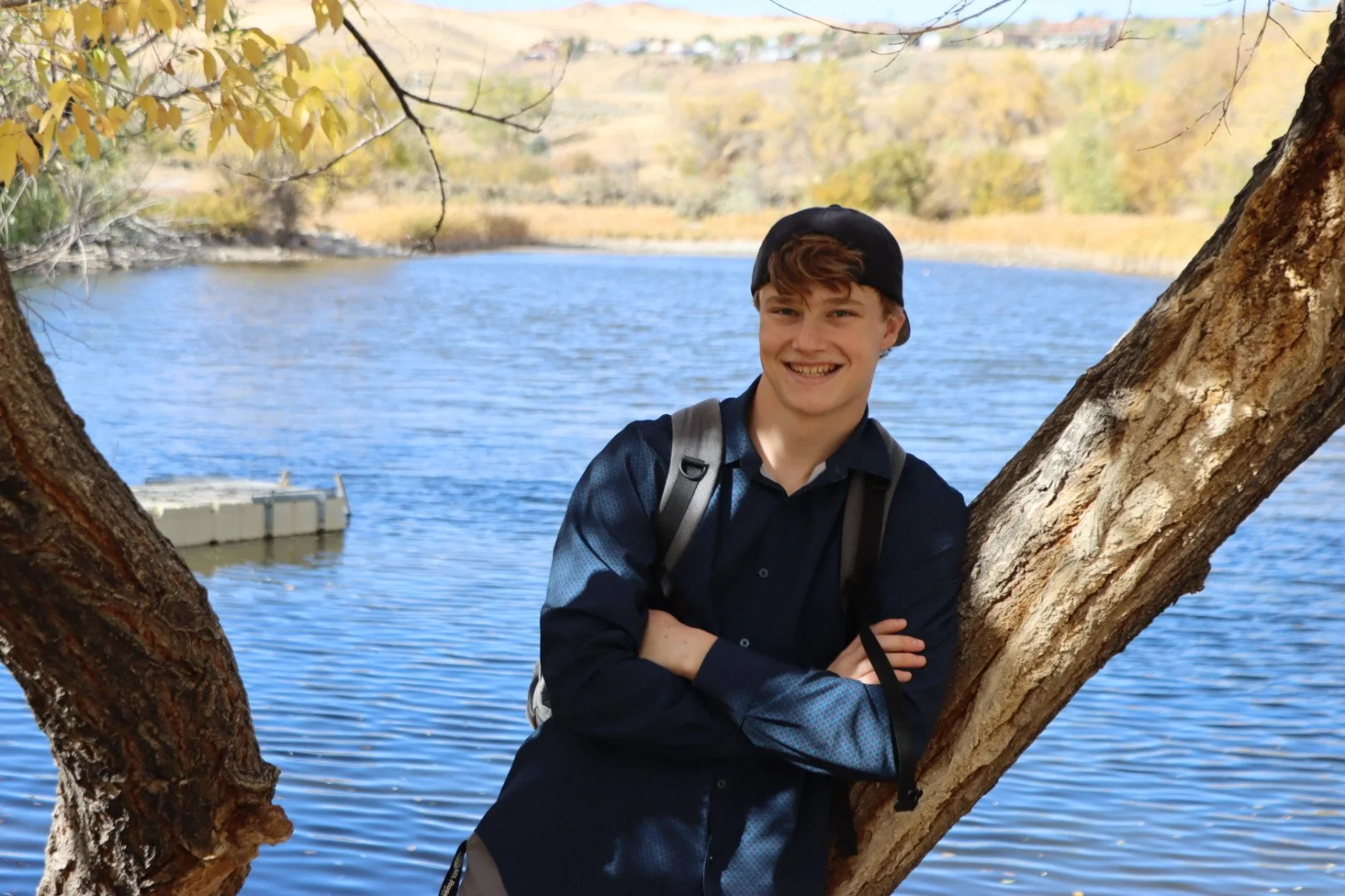 A smiling young man with light brown hair wearing a black cap, navy blue shirt, and a gray backpack, standing outdoors by a tree with arms crossed in front of a body of water with a small dock, and with autumn trees and hills in the background.