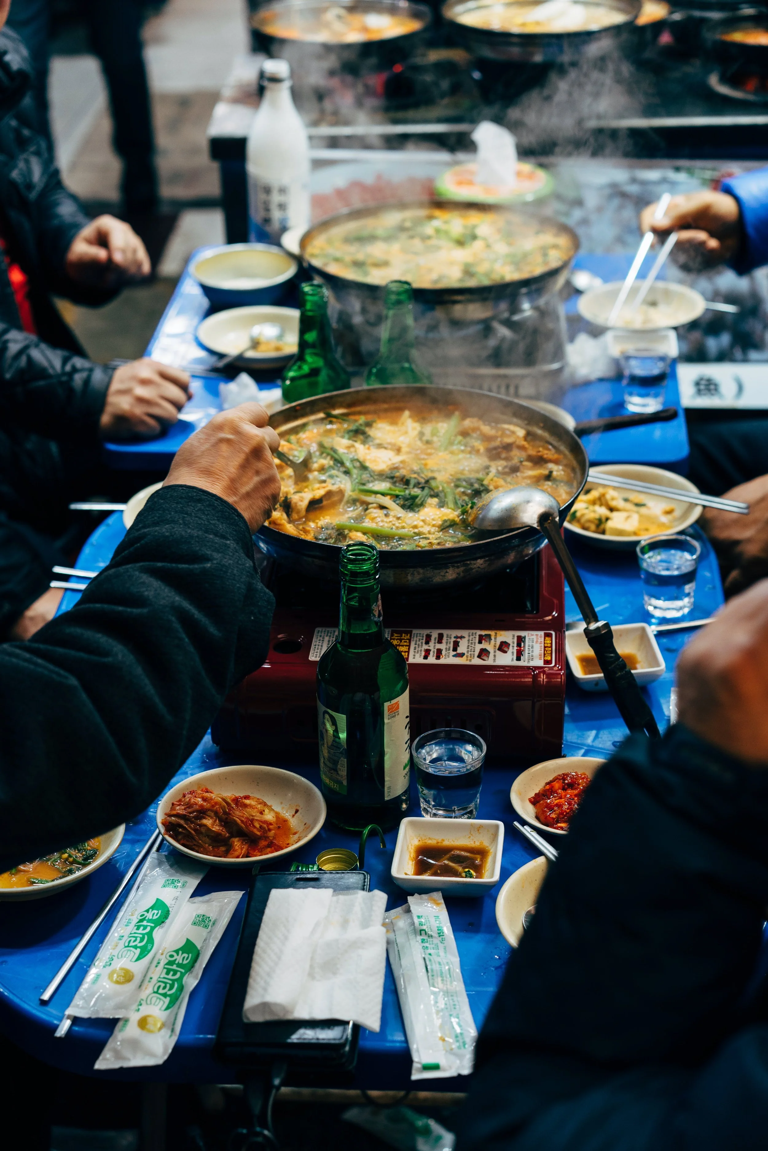 People sharing a hot pot meal at a restaurant, with steam rising from the pots and various side dishes and drinks on the table.