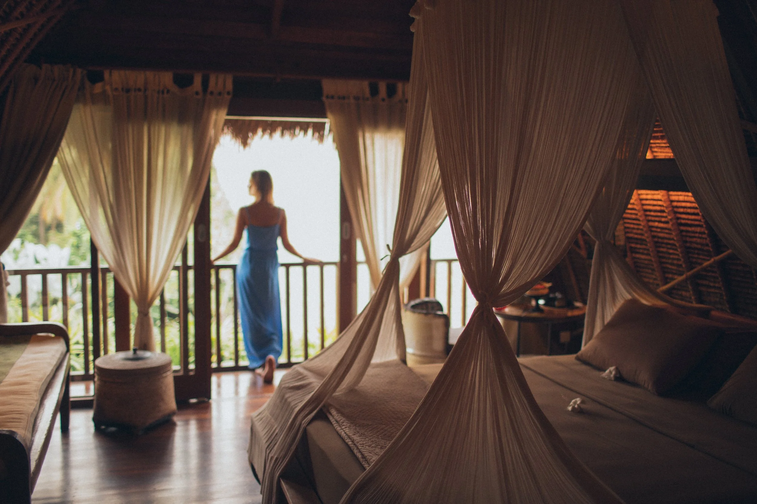 A woman in a blue dress standing on a balcony with curtains, overlooking a tropical landscape, inside a warmly lit room with a bed and wooden furnishings.