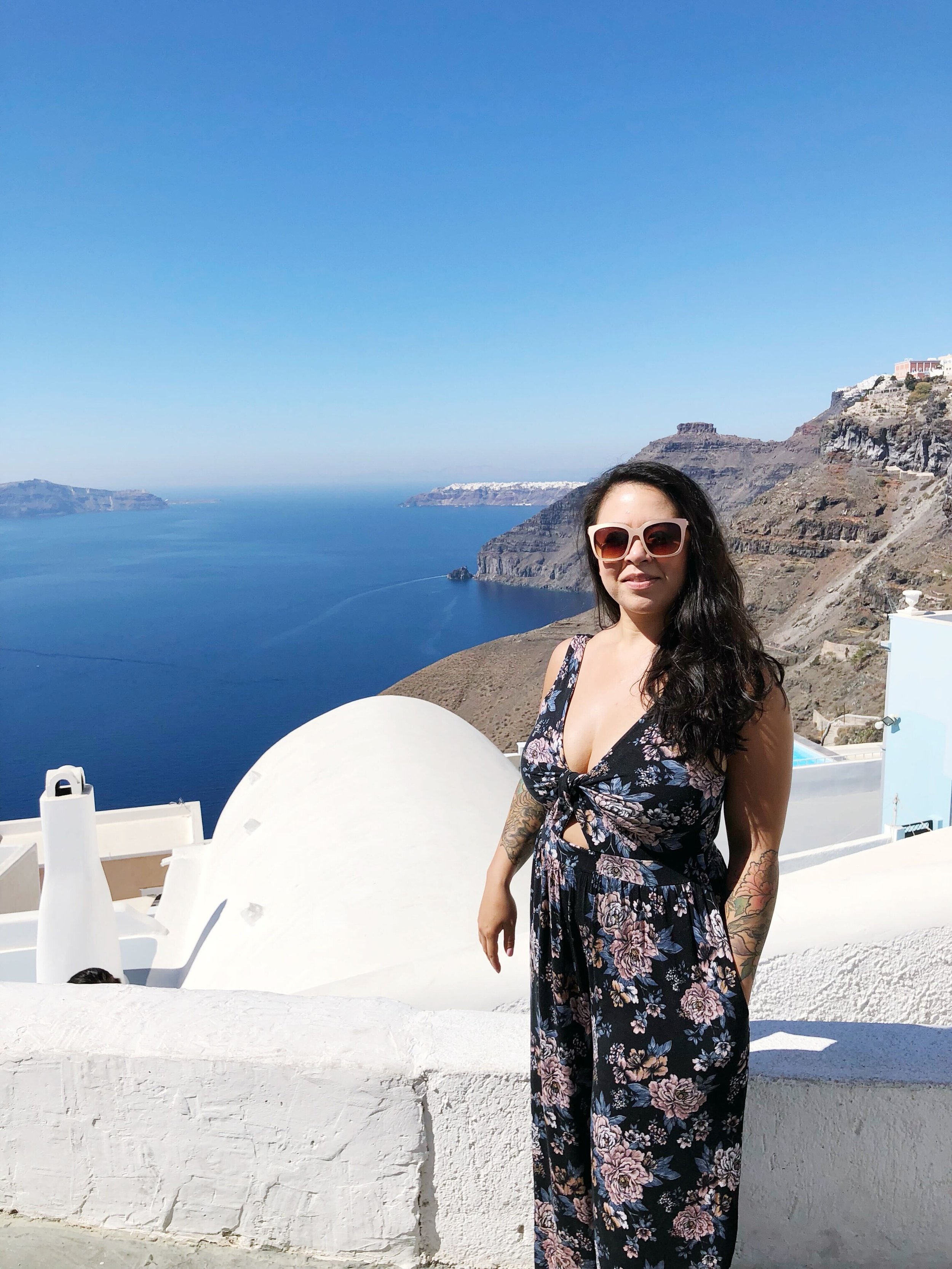 Woman with dark hair and tattoos on her arm wearing a floral jumpsuit and sunglasses, standing in front of a white building with a view of the blue sea and cliffs on the Greek island of Santorini.