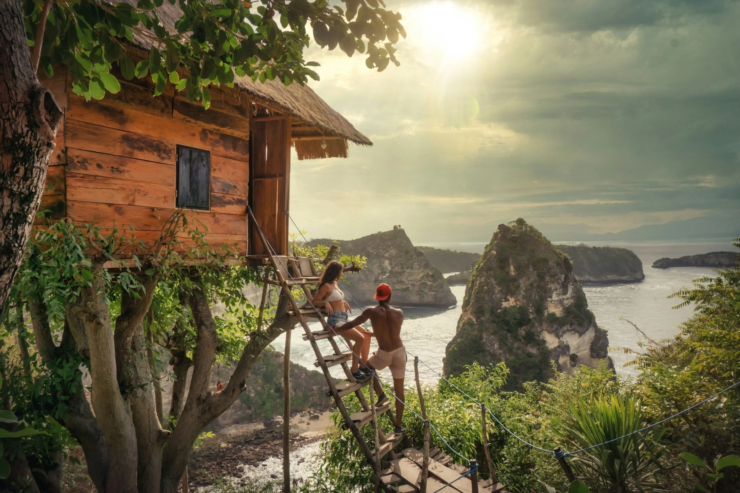 A wooden treehouse built into a large tree overlooking a coastal landscape with small islands and rocky formations, with two people interacting on the ladder leading up to the treehouse as the sun sets or rises in the sky.