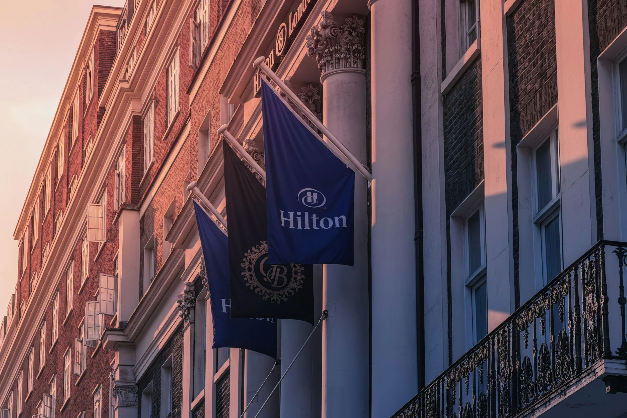 Exterior of a Hilton hotel with three blue and black flags displaying the Hilton logo hanging from the building's facade.