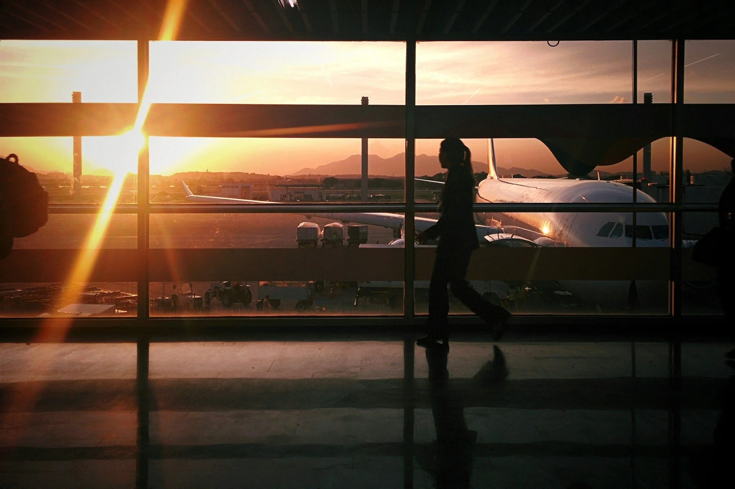 Silhouette of a person walking through an airport terminal at sunset, with an airplane visible outside the window and mountains in the distance.