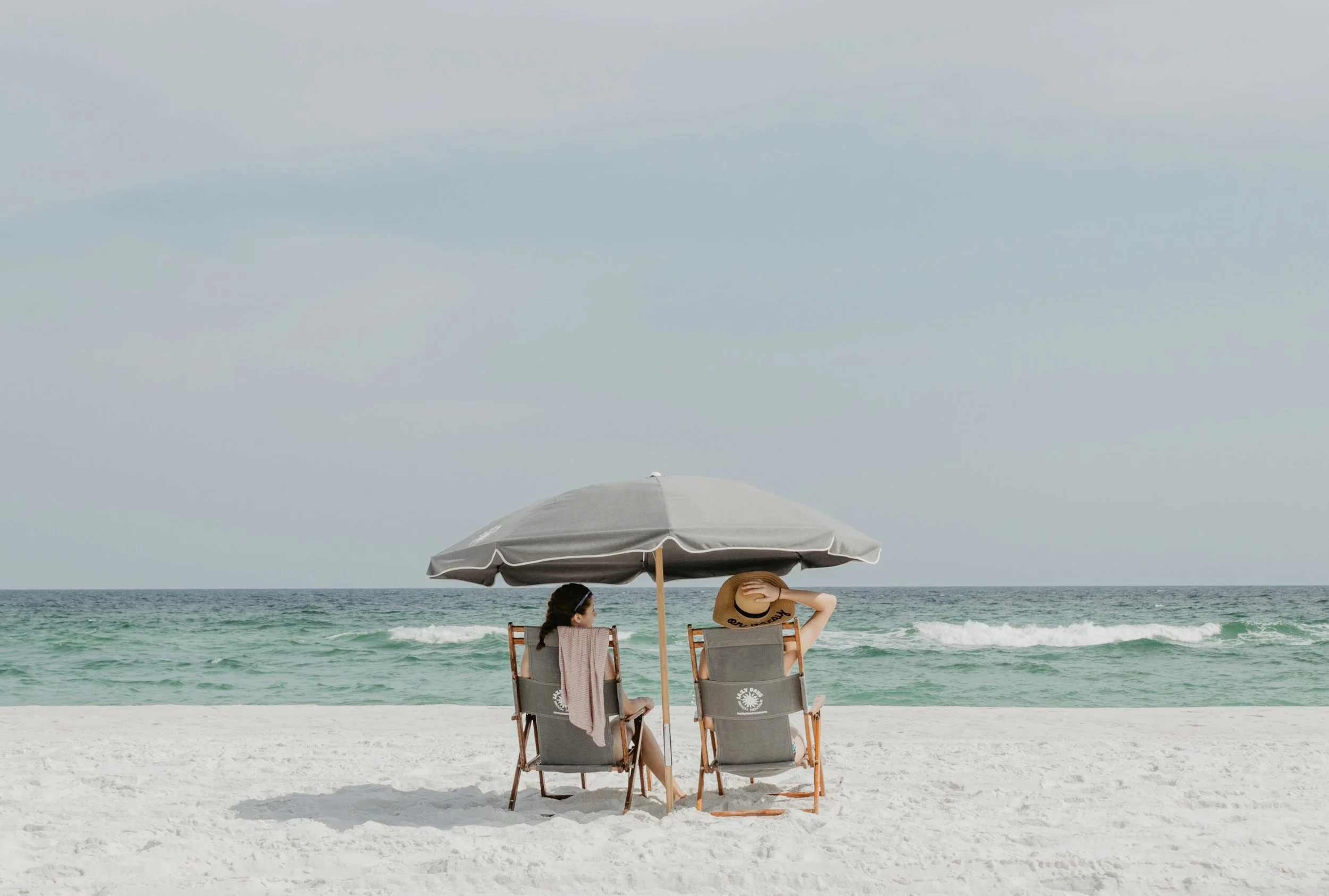 Two women sitting on beach chairs under a large beach umbrella on a sandy beach, facing the ocean with waves.