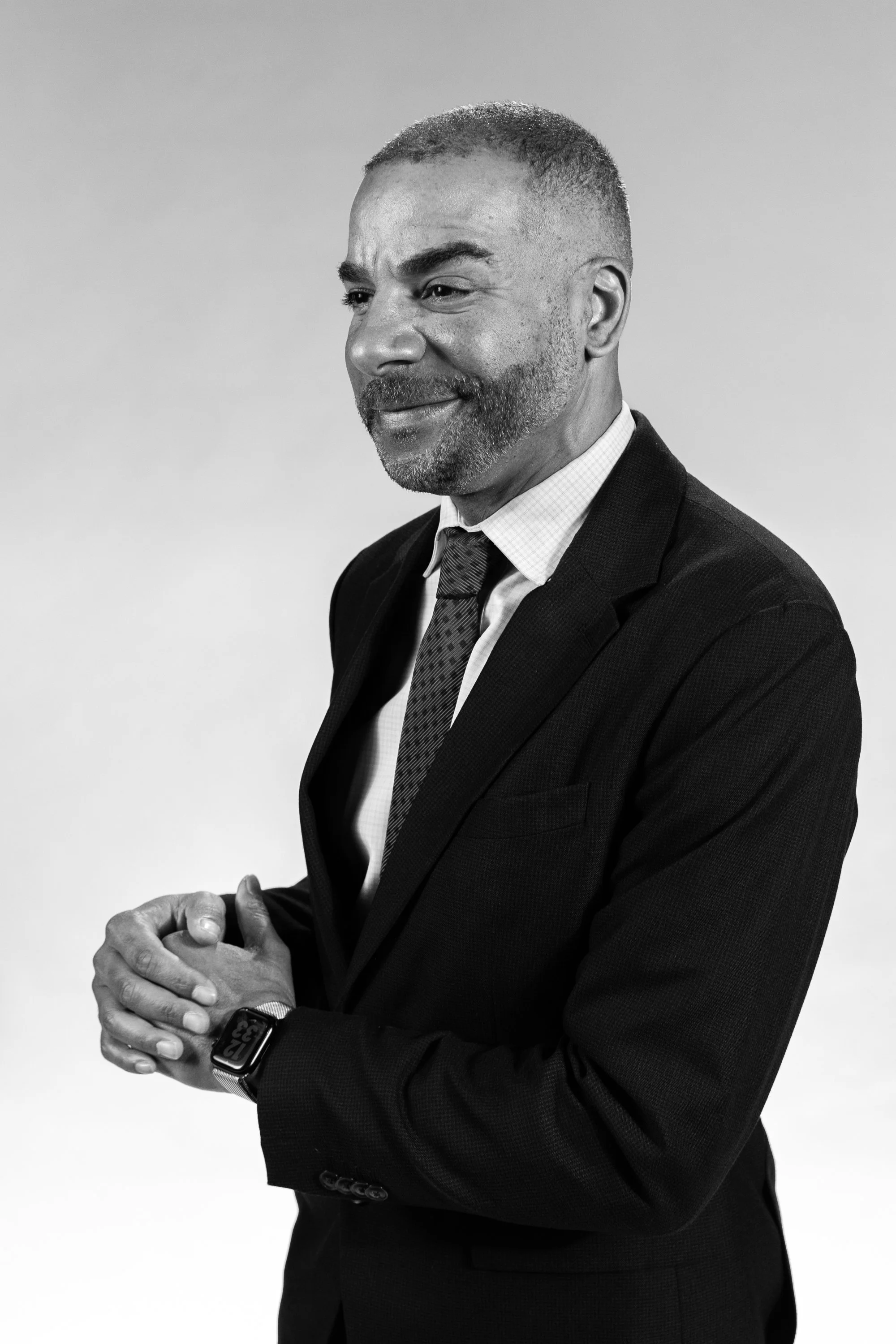 Black and white photo of a man wearing a suit, checking a smartwatch on his left wrist, with hands clasped together, against a plain background.