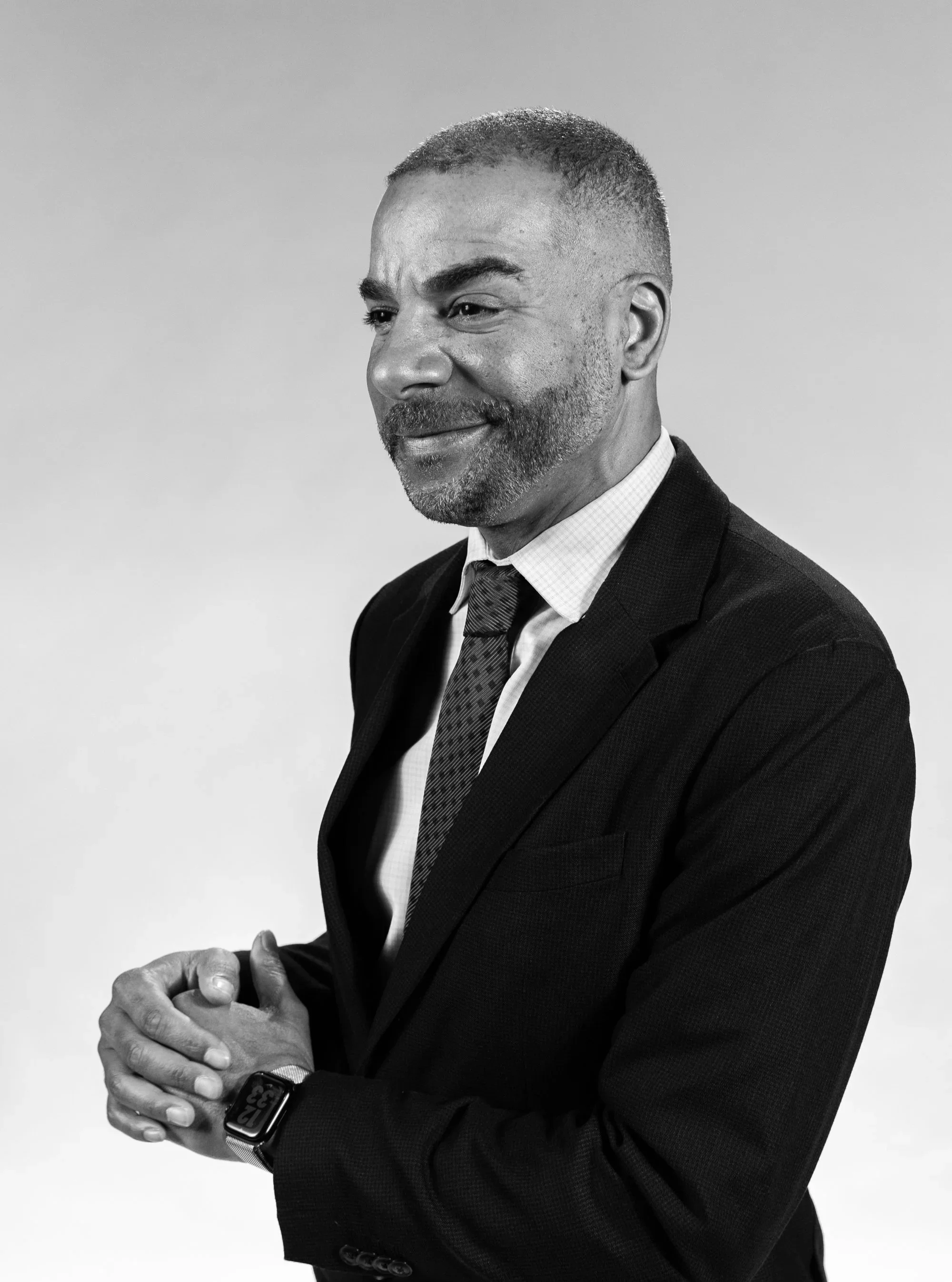 A black and white photo of a man dressed in a suit and tie, smiling with closed eyes, holding his hands clasped together in front of him, wearing a smartwatch on his wrist.