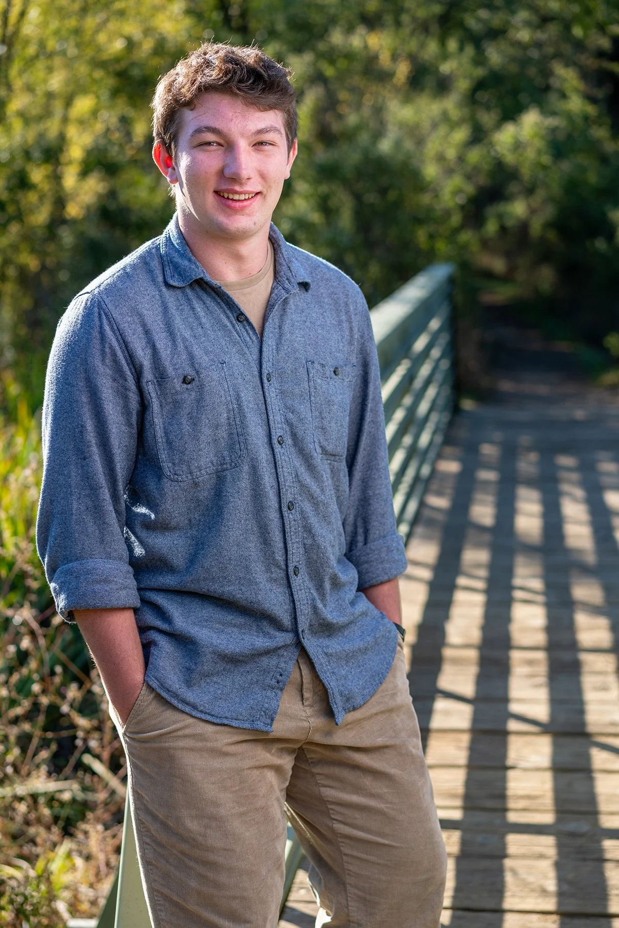 Young man smiling outdoors on a wooden bridge, wearing a denim shirt with rolled-up sleeves and khaki pants, with greenery in the background.