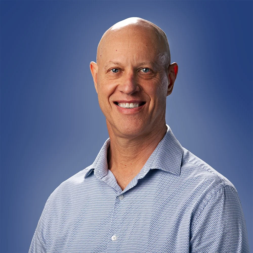 A bald man in a collared, patterned shirt smiling against a plain blue background.