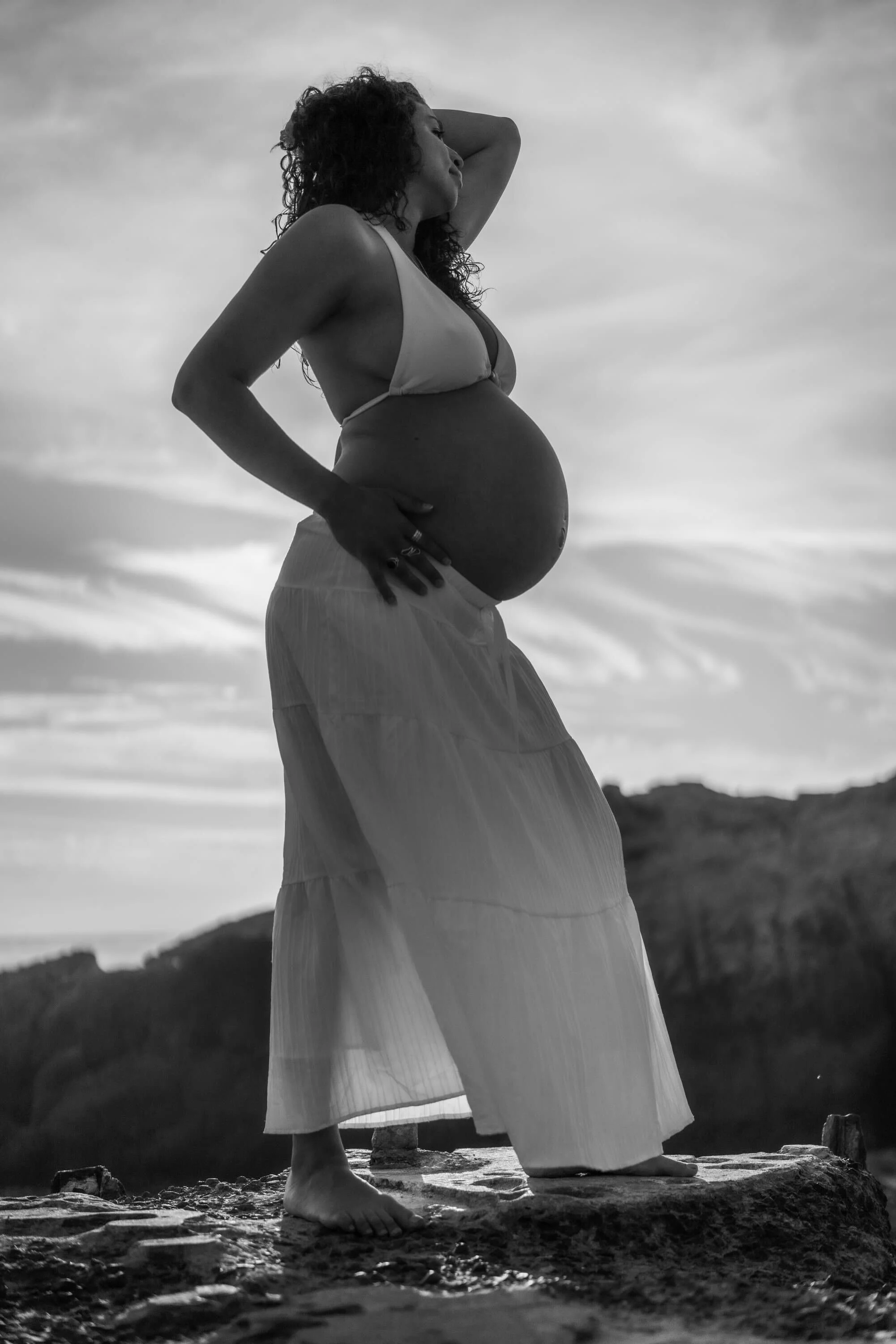 A woman pregnant and barefoot standing outdoors on a rock, wearing a bikini top and flowing skirt, with her left hand on her hip and her right hand resting on her head, gazing into the distance. The photo is in black and white with a cloudy sky in th
