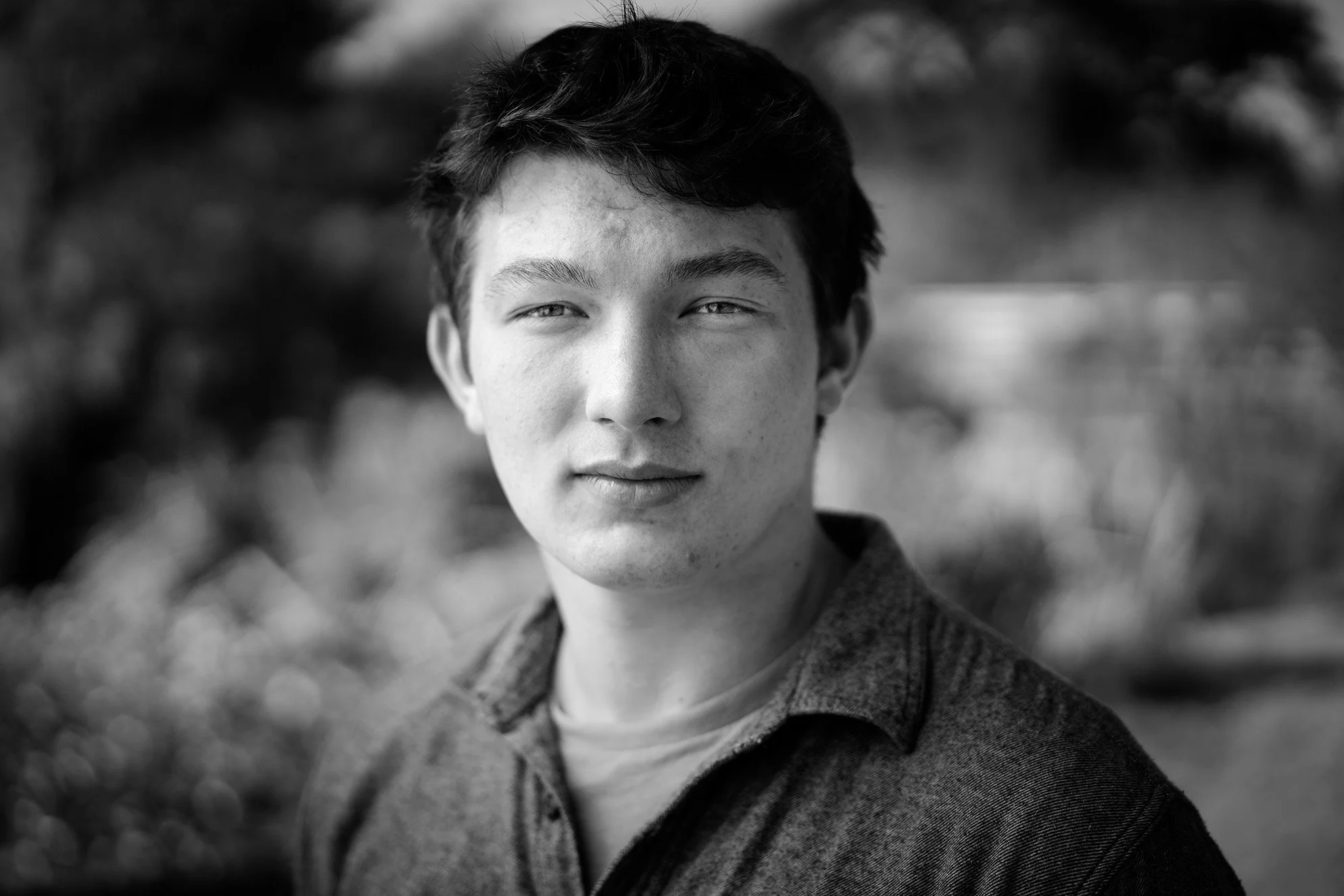 Black and white close-up portrait of a young man with short dark hair, wearing a dark jacket and light shirt, outdoors with blurred trees in background.
