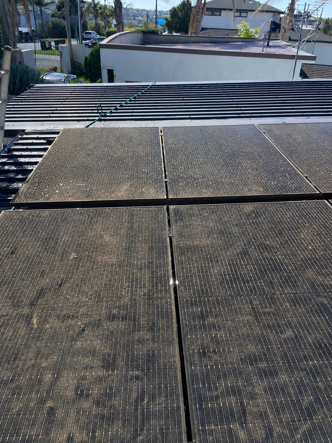 A view of solar panels on a rooftop, with dirt and dust on their surface, and neighboring residential buildings in the background.