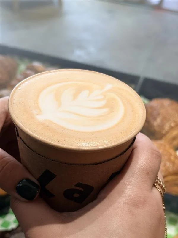 A hand holding a hot latte with latte art in the shape of a leaf or feather in a paper cup, inside a coffee shop with pastries in the background.