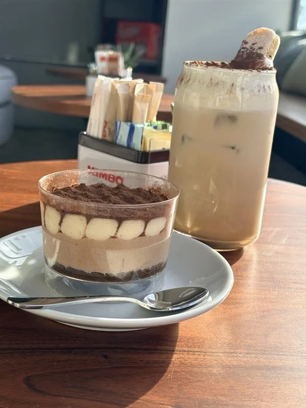 A glass of ice cream topped with cocoa powder on a white saucer with a spoon, and a large jar of iced coffee topped with whipped cream and a cookie, on a wooden table at a cafe.
