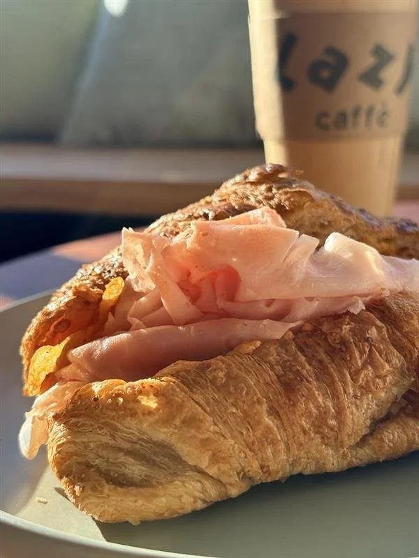A croissant sandwich with ham and turkey slices, served on a white plate. In the background, there is a paper cup with the cafe's logo, "Lazze cafe," likely containing coffee or a similar beverage.