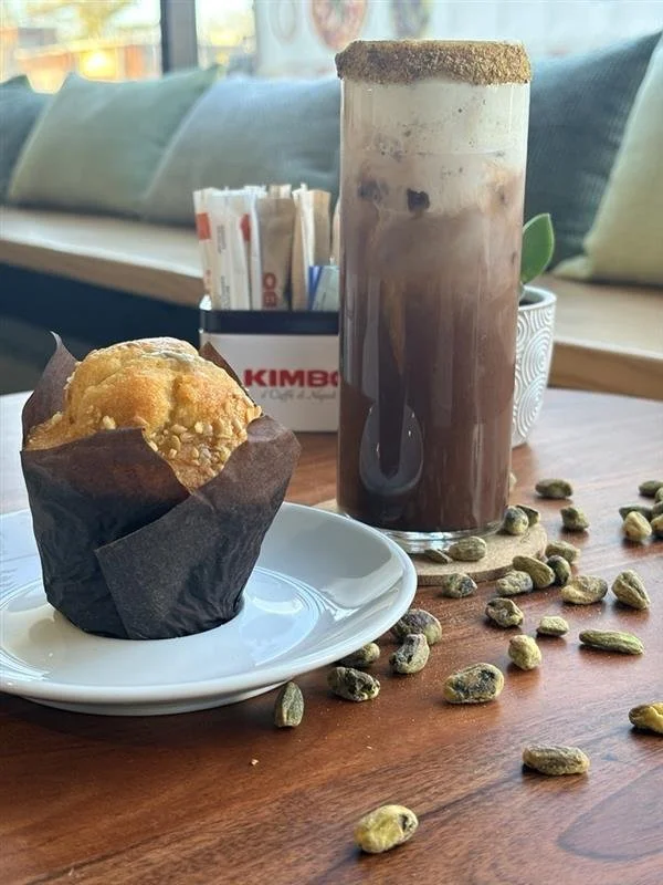 A muffin on a white plate and a glass of iced mocha coffee with whipped cream and a cookie on top, on a wooden table with scattered pumpkin seeds in a cafe.