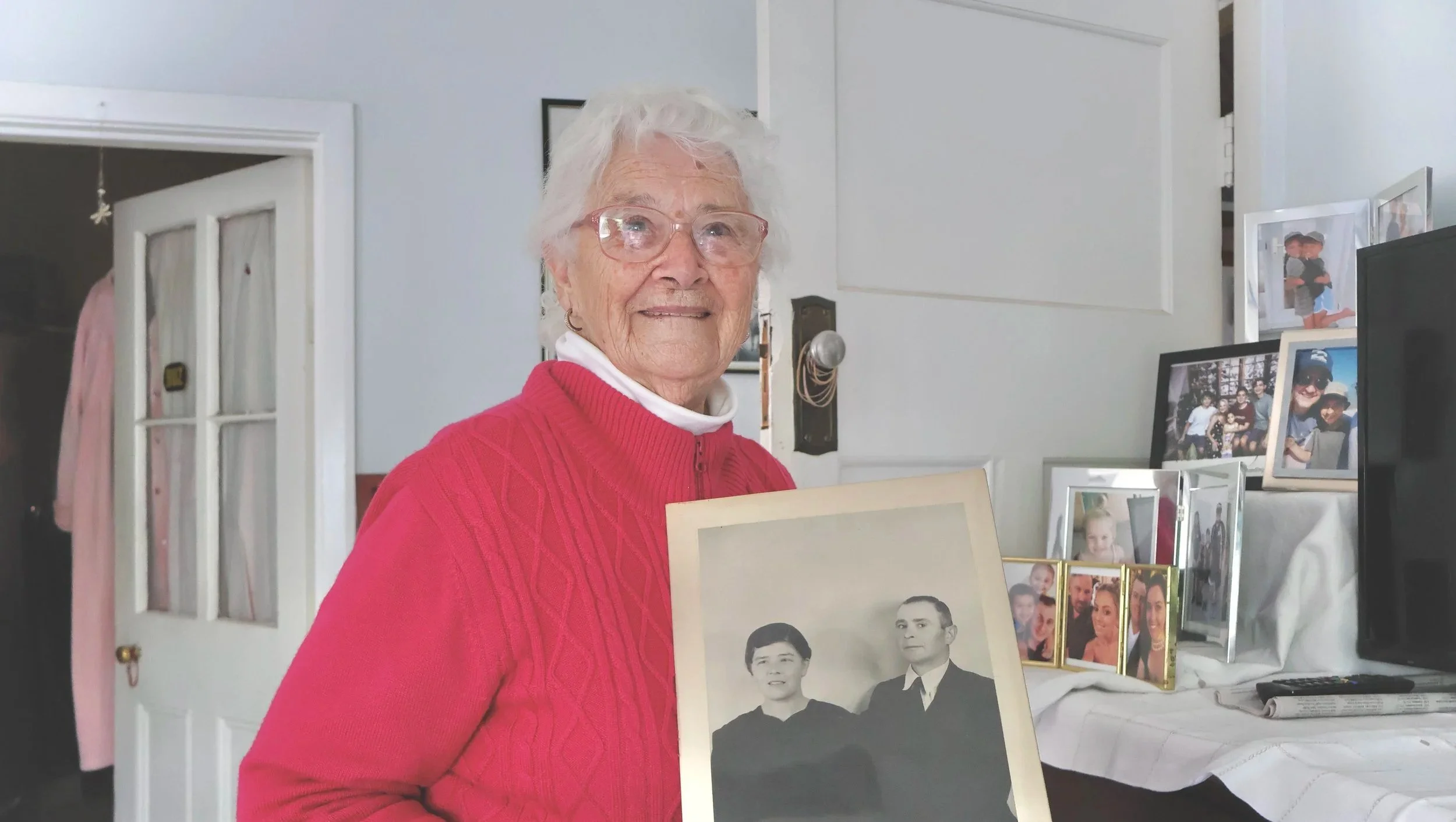 An elderly woman in Echuca with white hair and red glasses, wearing a red sweater, standing beside a display of framed photographs and holding a black-and-white portrait of her parents.