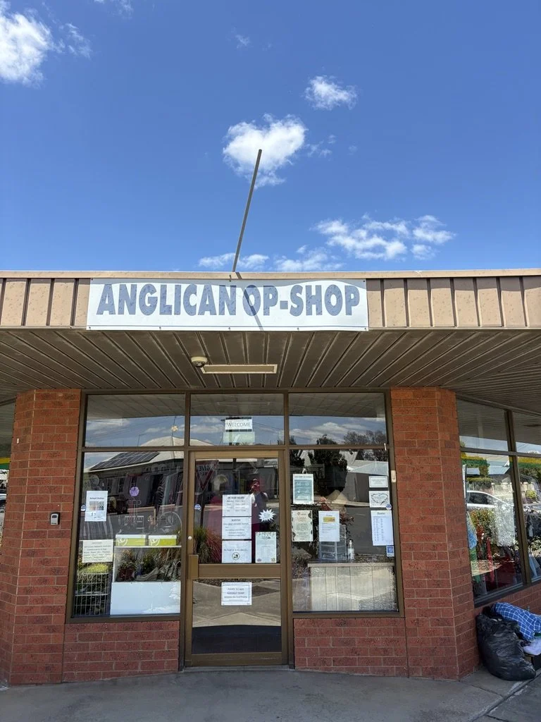 Front view of an Anglican Op Shop storefront with a glass door and large windows, brick columns on either side, and signage above the entrance, under a partly cloudy sky.