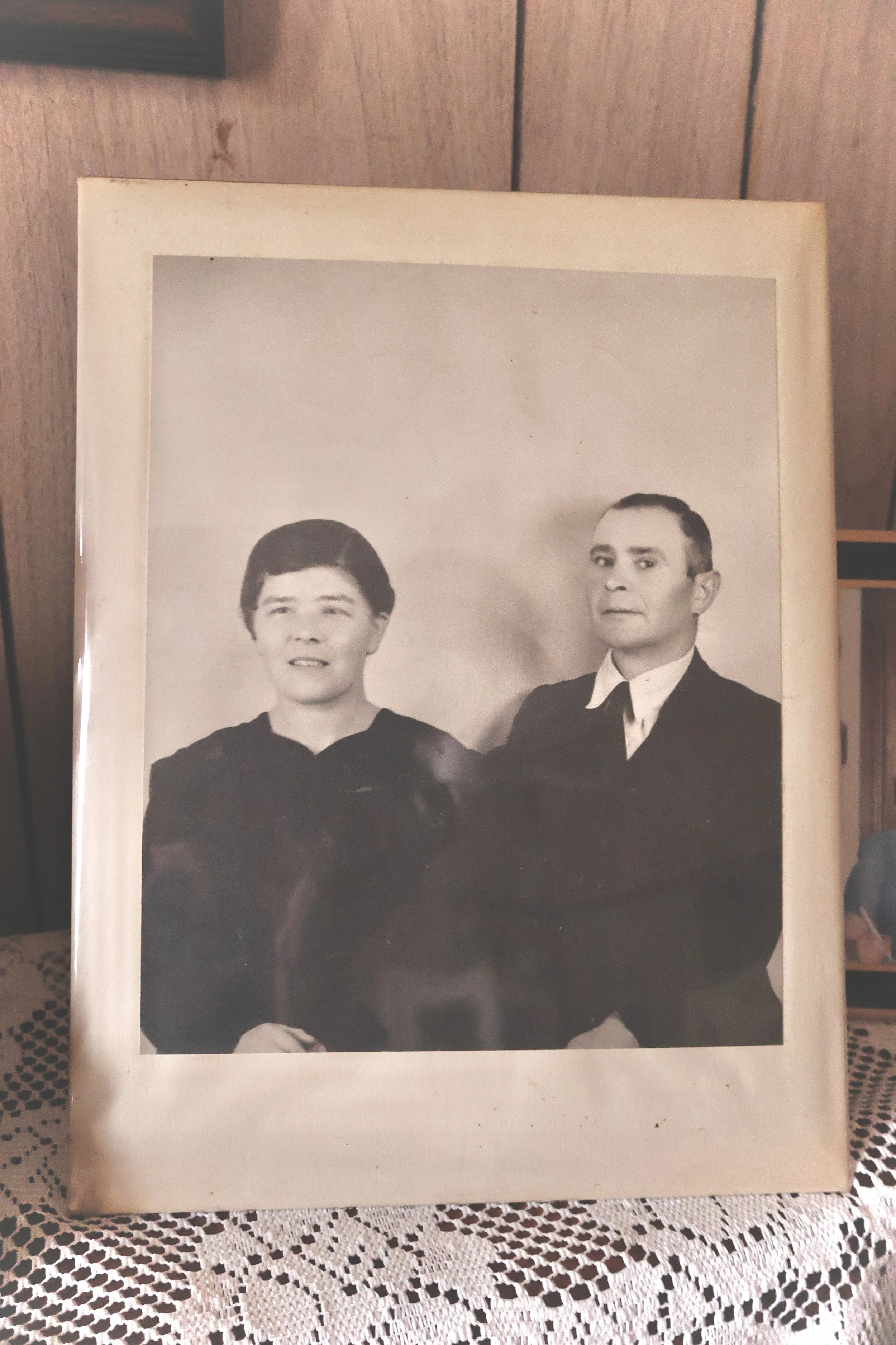 Black and white portrait photo of two men in formal attire, seated with hands crossed, on a lace tablecloth.