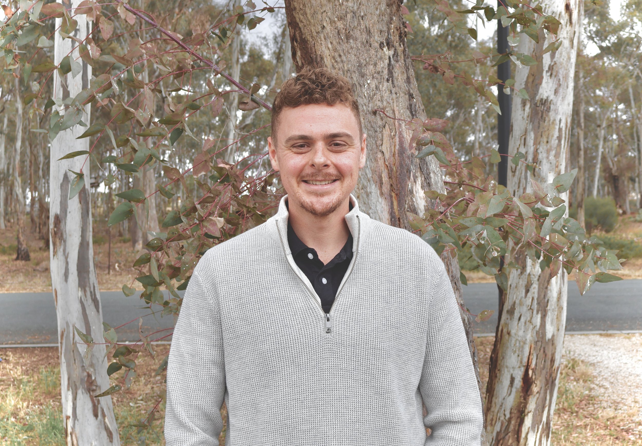 A smiling young man with short curly hair and a beard, wearing a light gray zip-up sweater, standing outdoors in front of a tree with peeling bark and green leaves, in a park or wooded area.