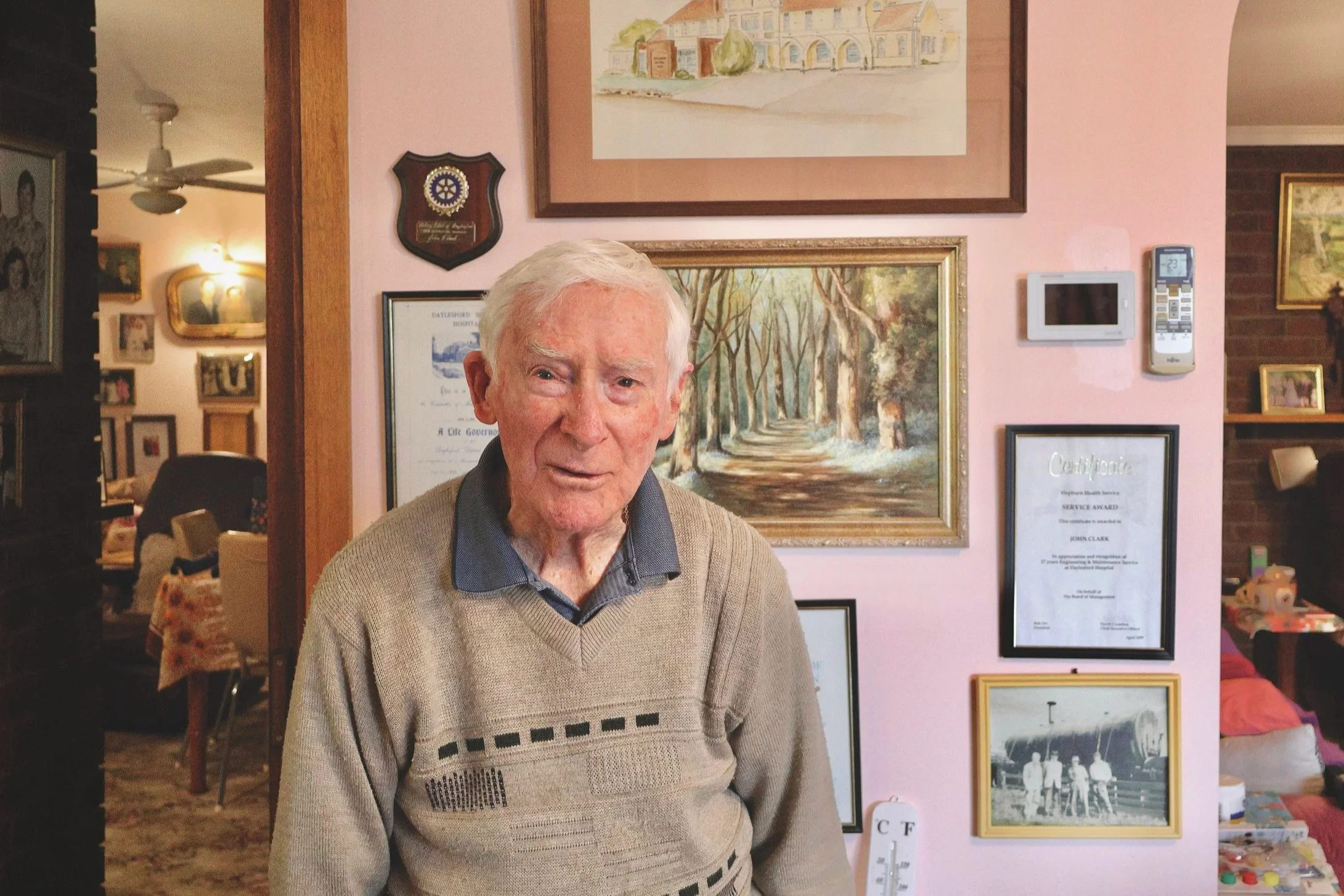 An elderly man in Daylesford with white hair and a beige sweater standing indoors with framed pictures and artwork on the pink wall behind him.