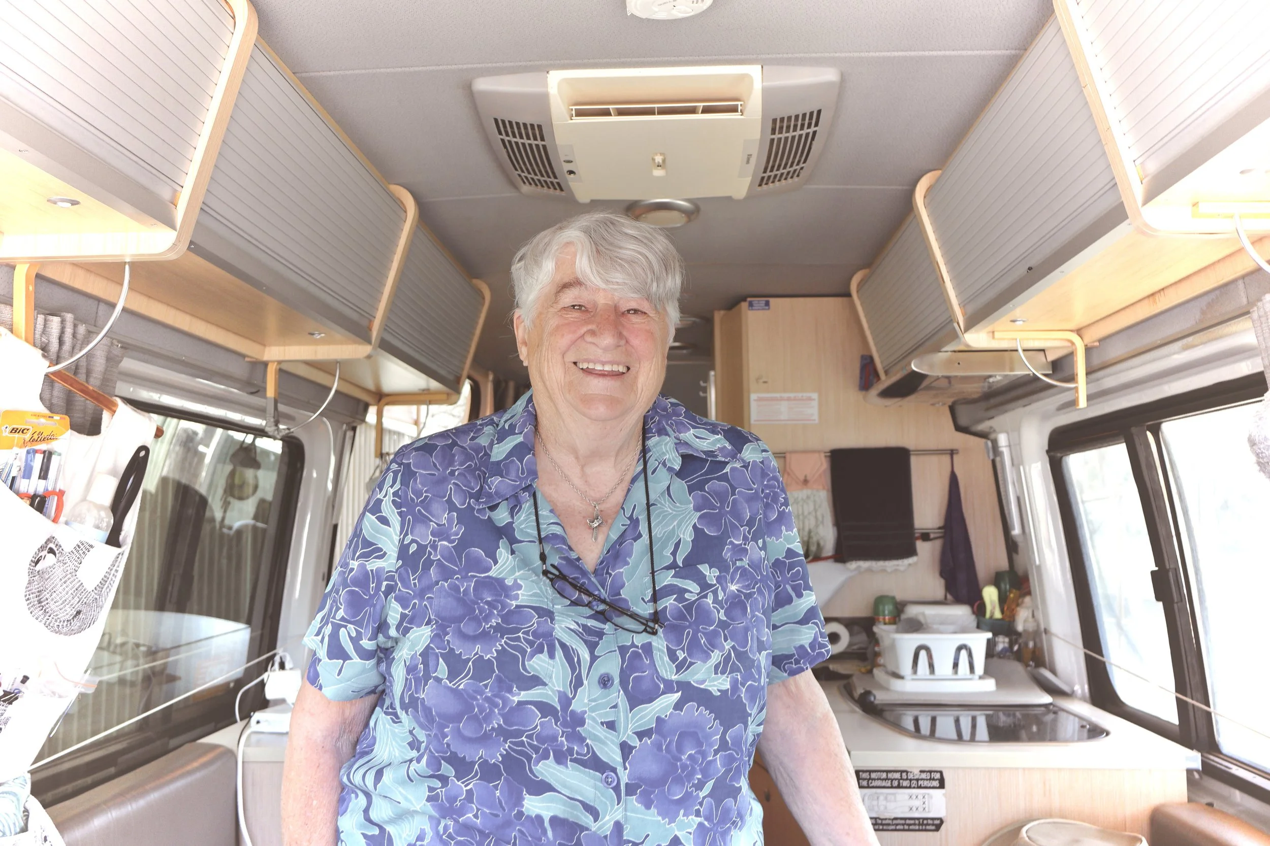 The image shows an elderly man with gray hair smiling inside a camper van or RV. He is wearing a blue Hawaiian shirt featuring a floral pattern, glasses hanging around his neck, and a necklace. The interior of the camper has light wood cabinets, a kitchenette, and various household items.