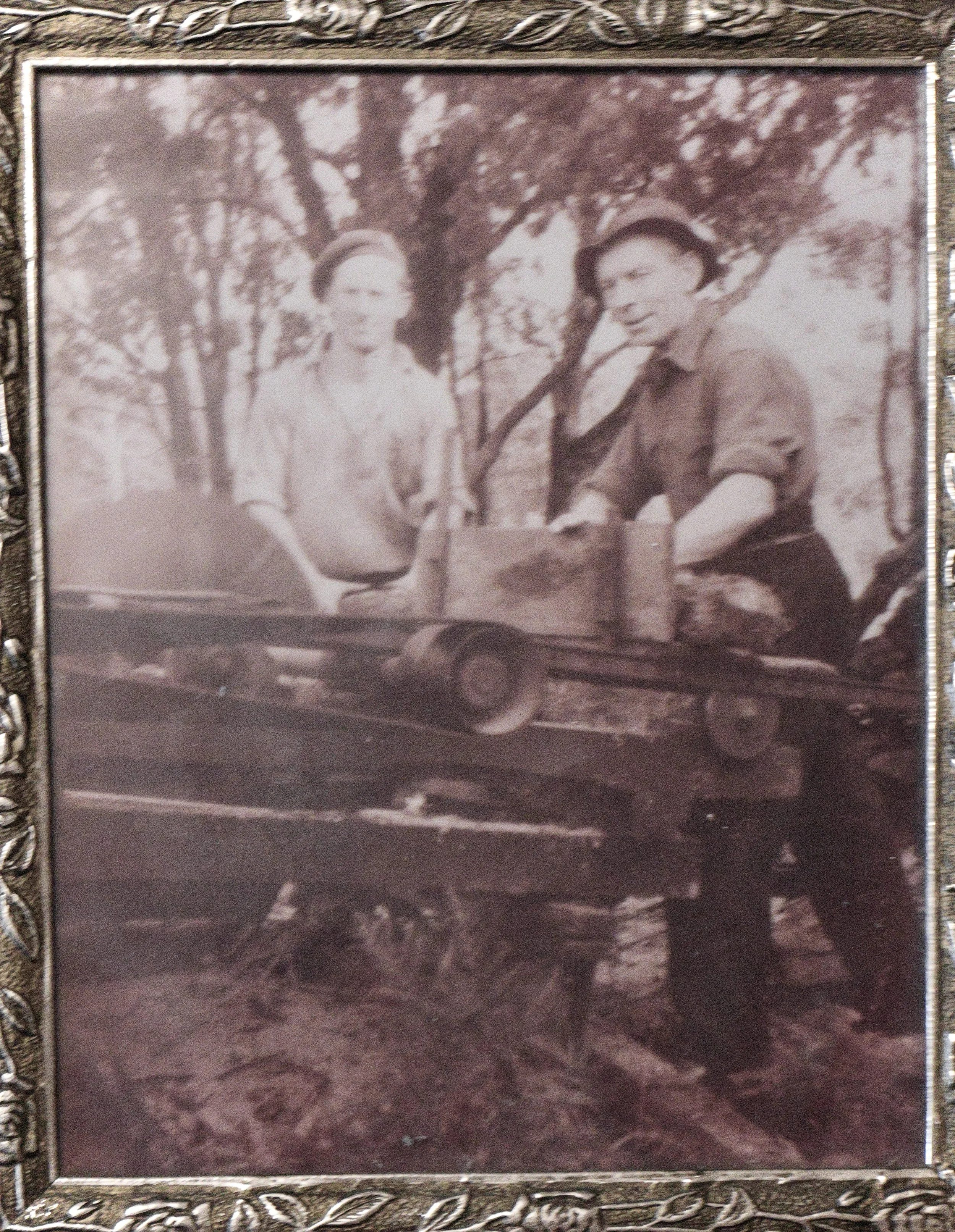 An old black and white photograph of a man and a woman sitting outside on a wooden bench in front of trees. The woman is wearing a blouse and the man is wearing a hat and a shirt. They are looking at the camera.