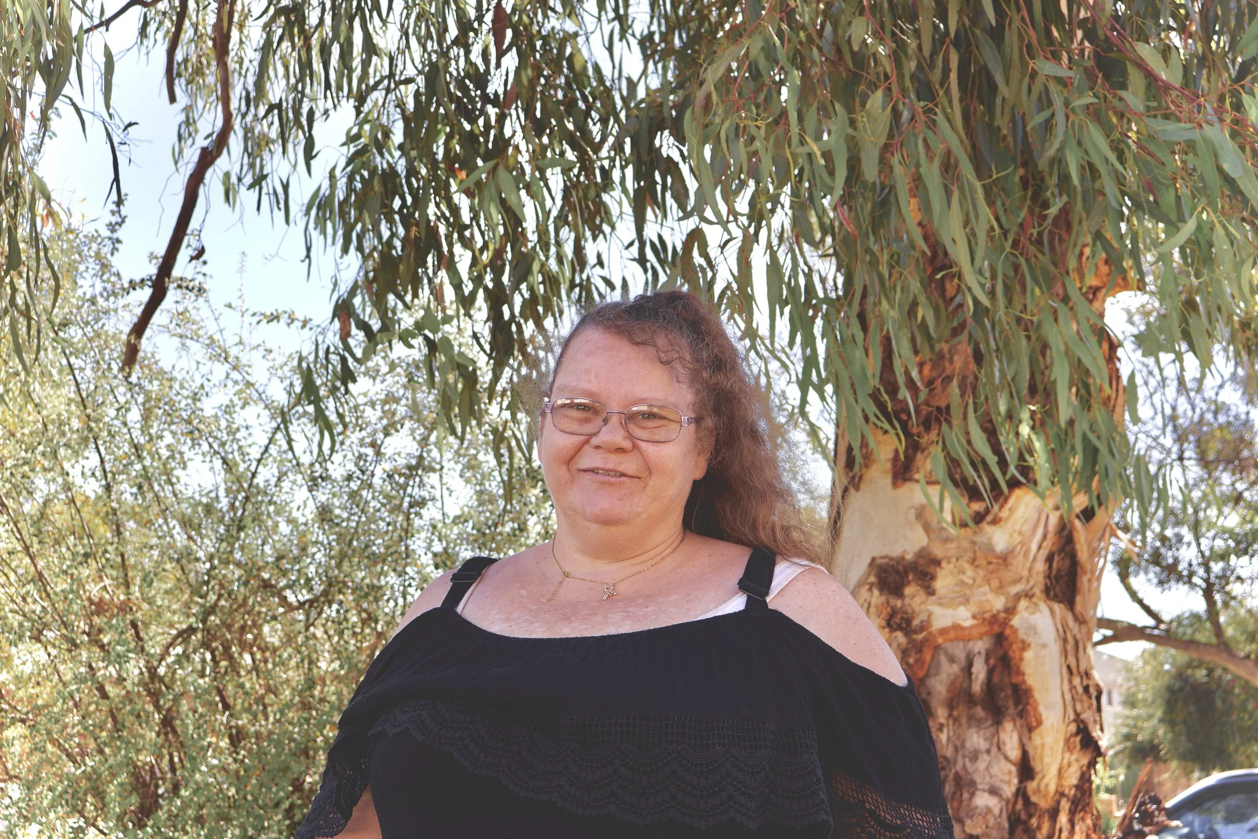 A woman with curly hair and glasses standing outdoors in front of a large tree with hanging branches and leaves, smiling at the camera.