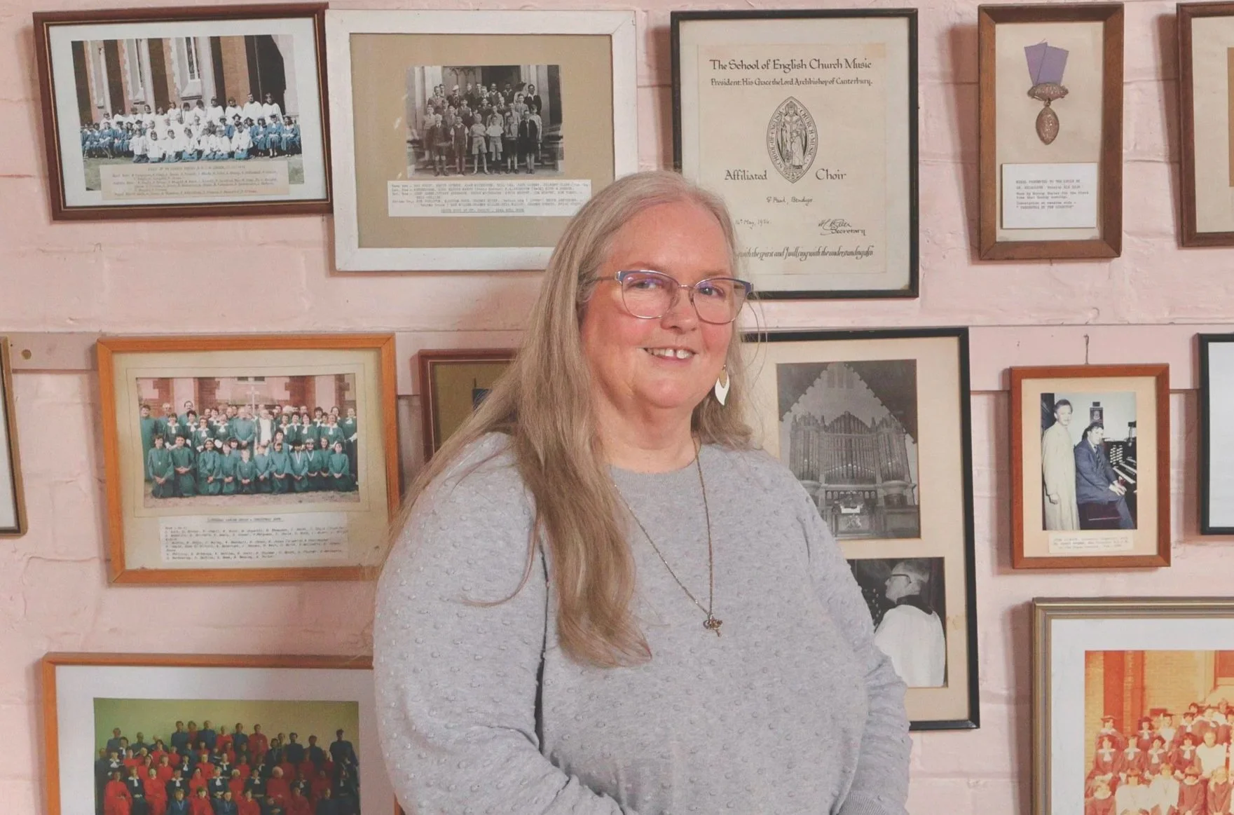 A woman with long blonde hair, glasses, and earrings stands in front of a pink brick wall decorated with framed photographs and certificates from St Paul's Cathedral Choir, Bendigo. She is smiling and wearing a light gray sweater.