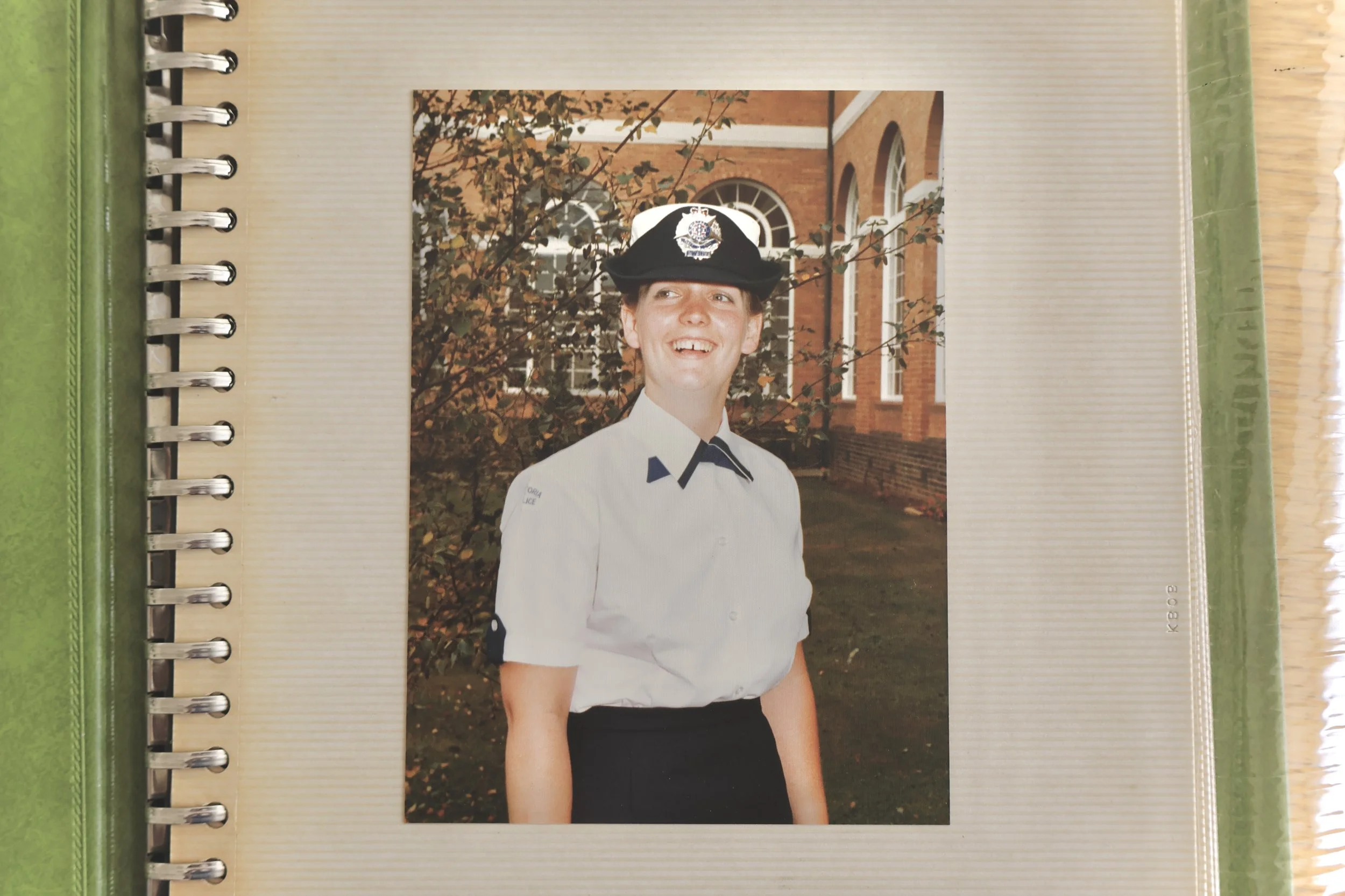 A young man in a police uniform with a hat, smiling, standing outdoors in front of a brick building with arched windows, surrounded by trees with autumn leaves.